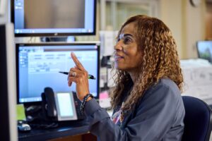 A woman with curly hair is sitting at a desk, pointing at a computer screen. She holds a pen in her other hand and is surrounded by office equipment and monitors. She appears focused and engaged.