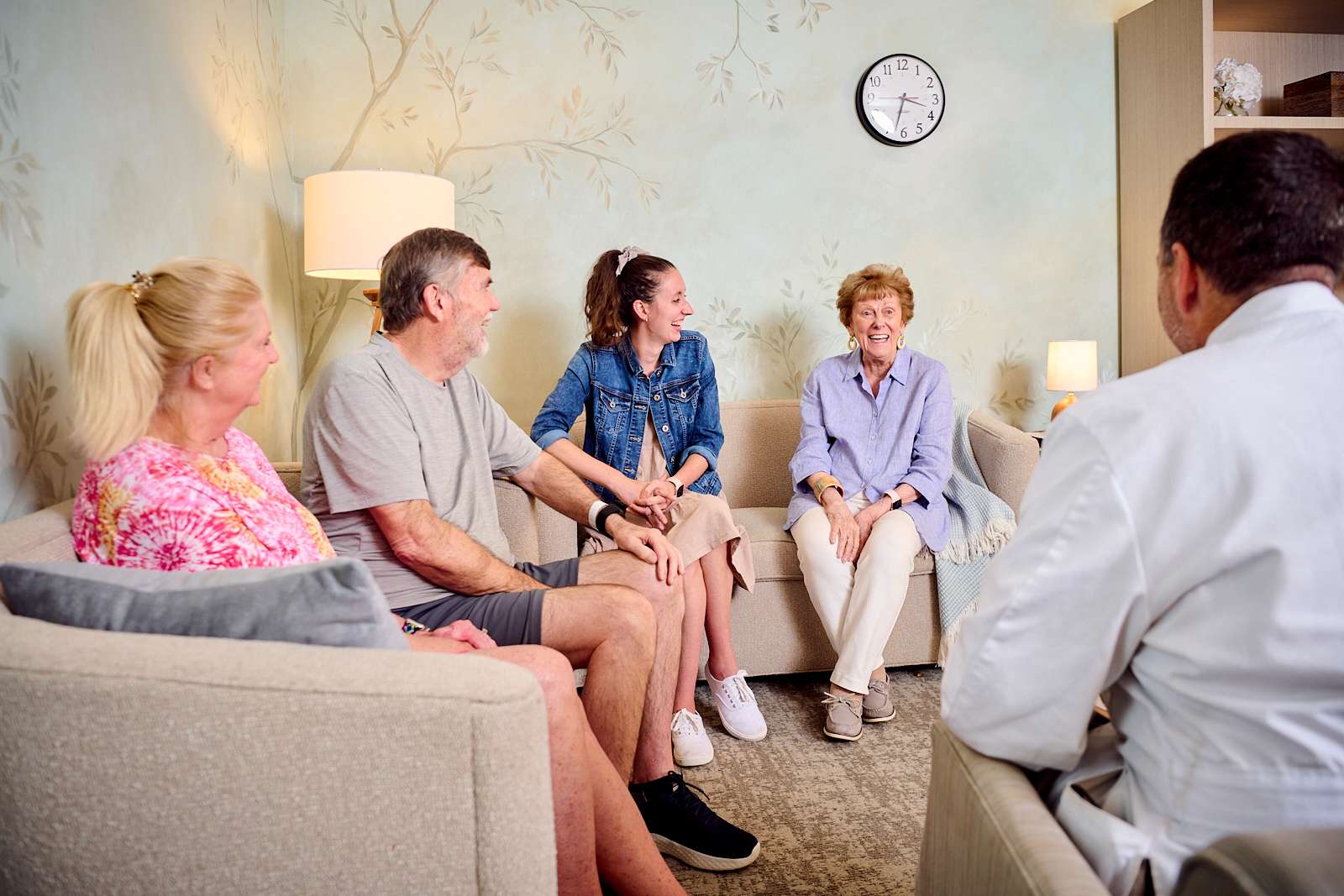 In a well-lit waiting room with neutral decor, five people, including medical personnel, patient and family, sit engaged in a discussion. They appear smiling and relaxed. A lamp and clock adorn the wall, while bookshelves in the background.
