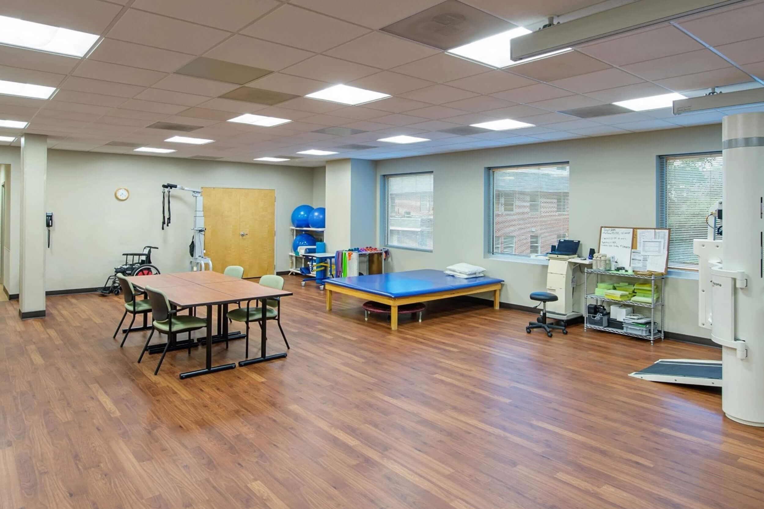 A physical therapy room with wooden flooring, a treatment table, exercise balls, chairs around a table, and various therapy equipment. Large windows let in natural light, and storage shelves line the walls.