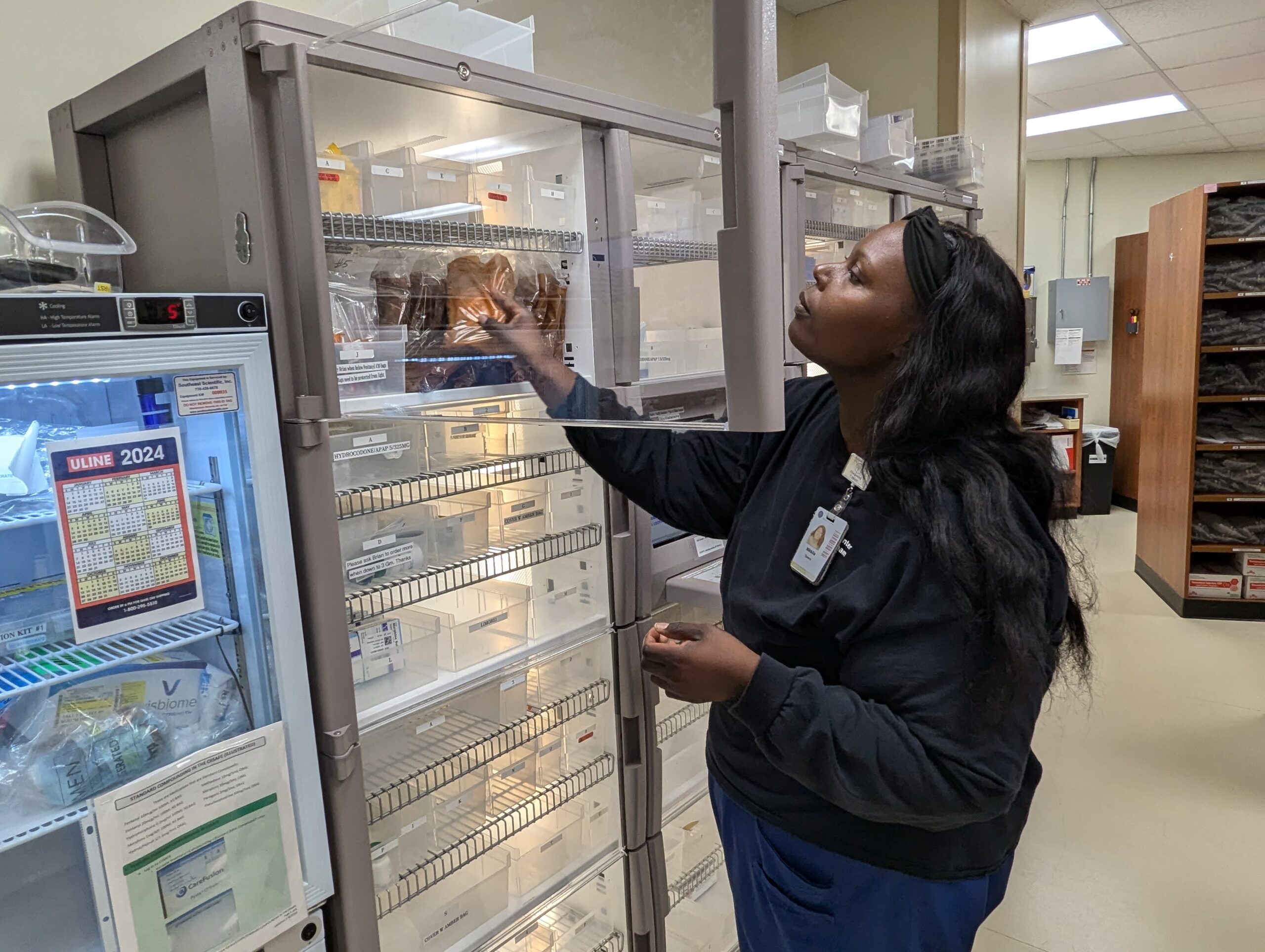A woman in a medical uniform organizes supplies in a well-lit storage room. She places items on a shelf while standing next to a cabinet with various compartments.