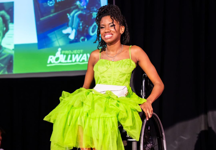 A young lady in a vibrant green dress sits in a wheelchair on stage, smiling confidently. An audience applauds in front. The background displays photos on a screen.