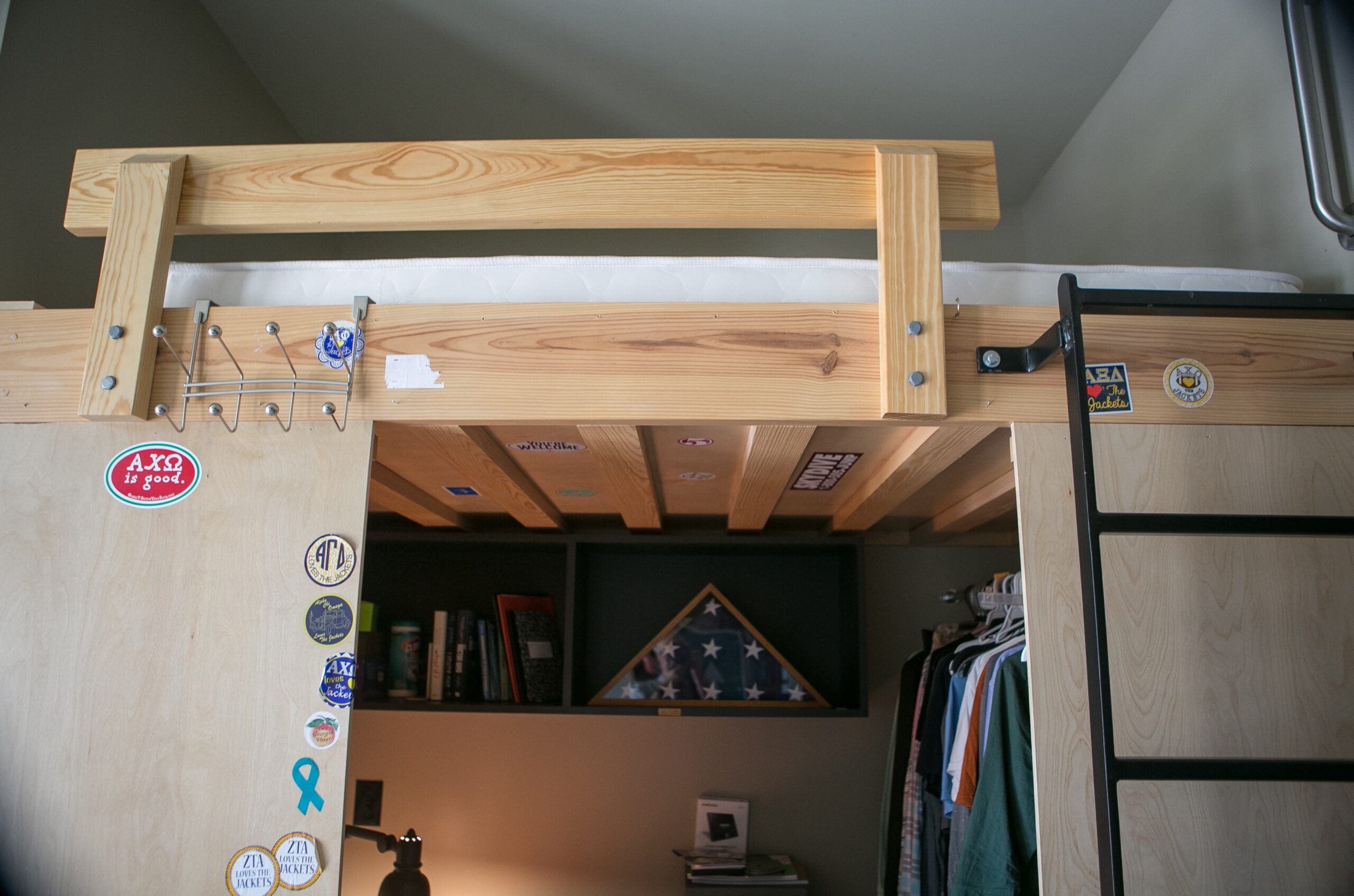 A wooden loft bed with a ladder attached. Below the bed, a closet with clothes and some shelves. The bed frame is adorned with various stickers. A folded American flag is displayed on one shelf.