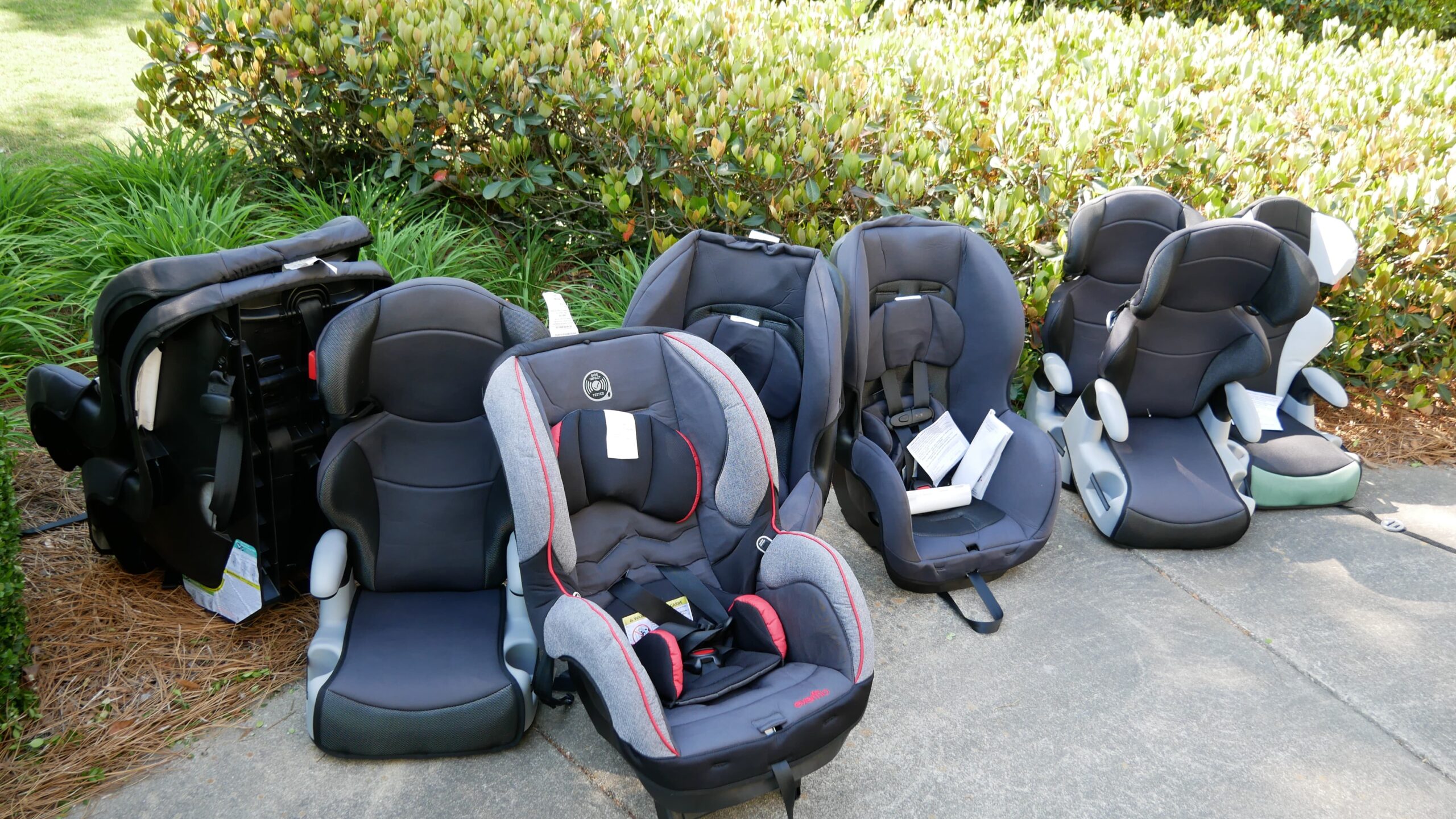 Several child car seats are lined up on a sidewalk near a grassy area and bushes. The seats vary in design and color, with some being black and others featuring gray fabric with red accents. They're arranged from left to right.
