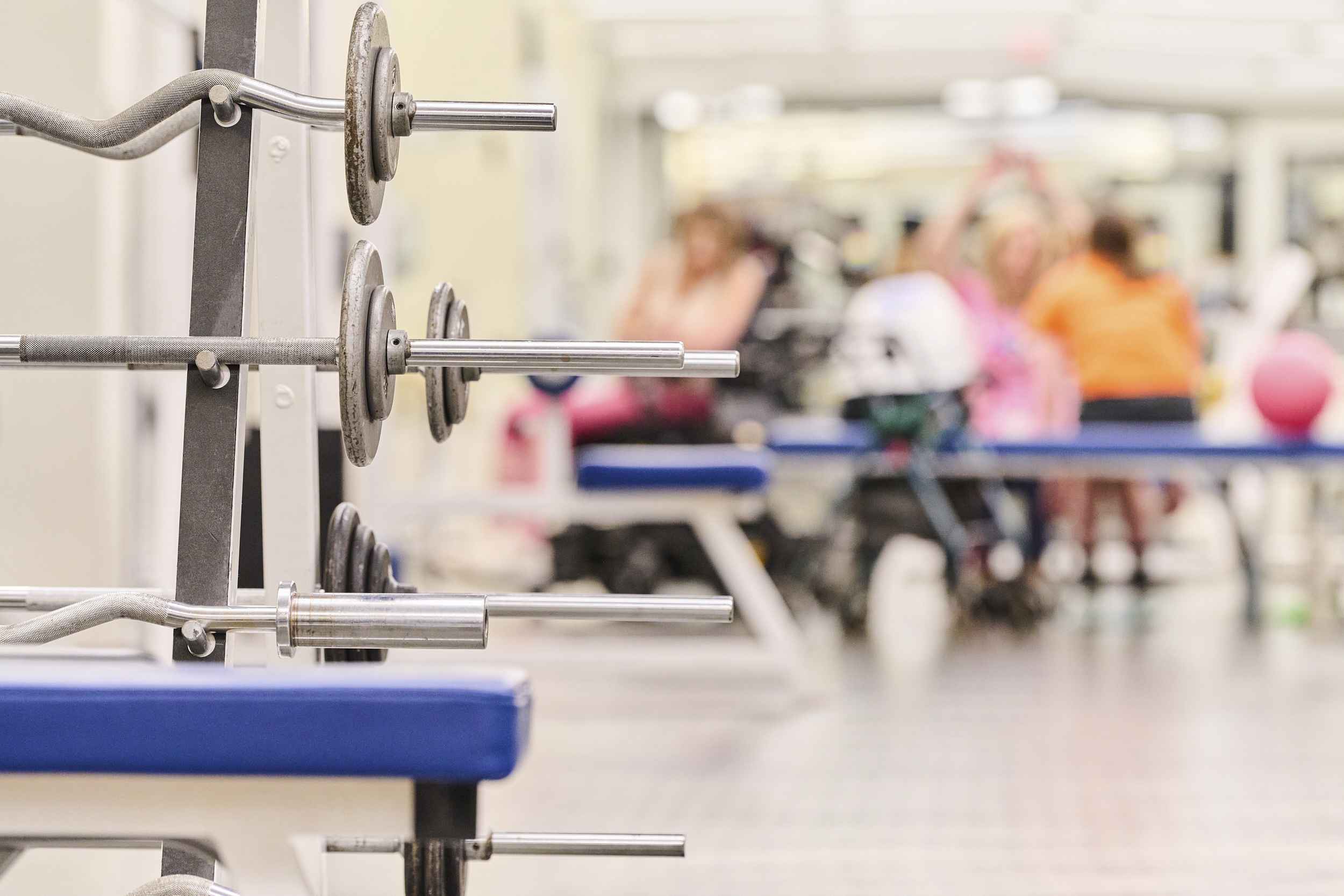 Barbells are stacked on a rack in the foreground of a gym. In the blurred background, people are gathered near a bench, engaging in activities. The setting appears spacious and bright.
