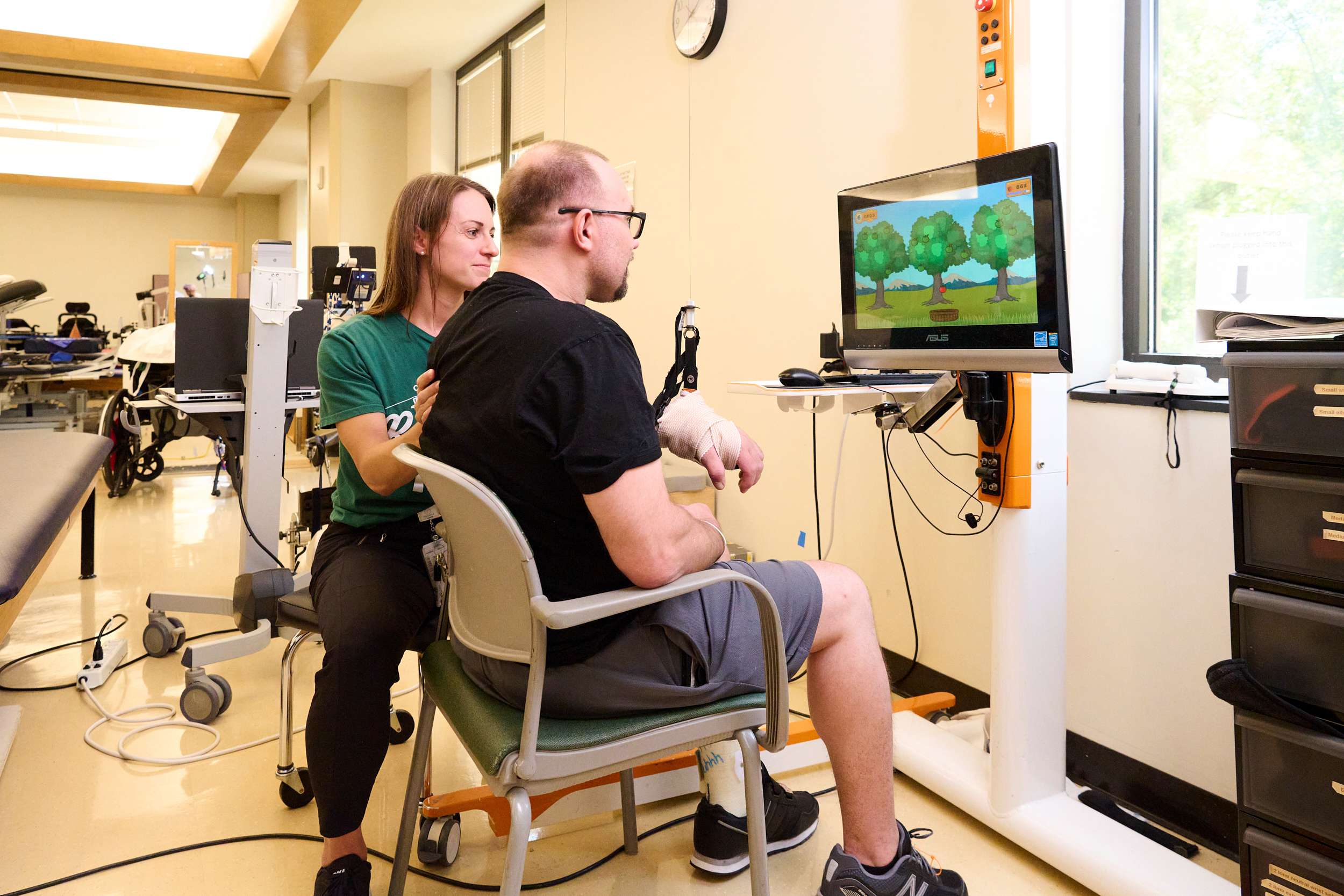 A therapist assists a man sitting in front of a screen during his physical therapy session. He is focused on an interactive program displaying trees. The room is filled with various rehabilitation tools, enhancing his recovery journey.