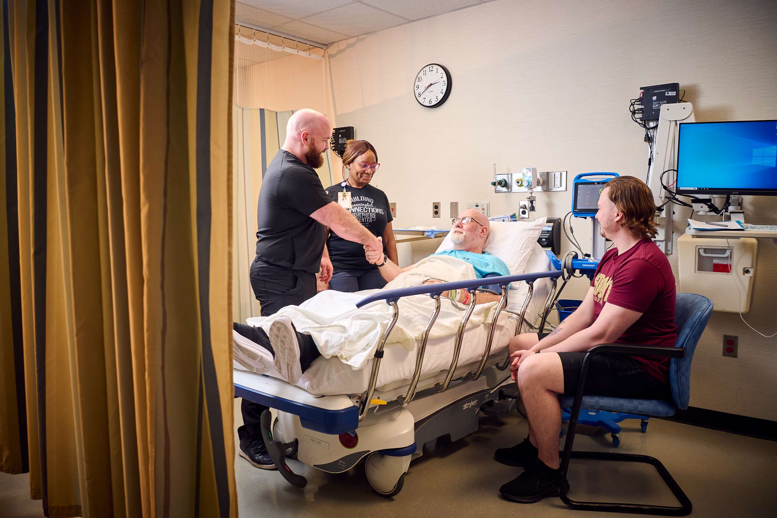A patient lies in a hospital bed with a man shaking hands and a woman standing beside him. Another young man is seated, observing the interaction. Medical equipment and a wall clock are visible in the background.