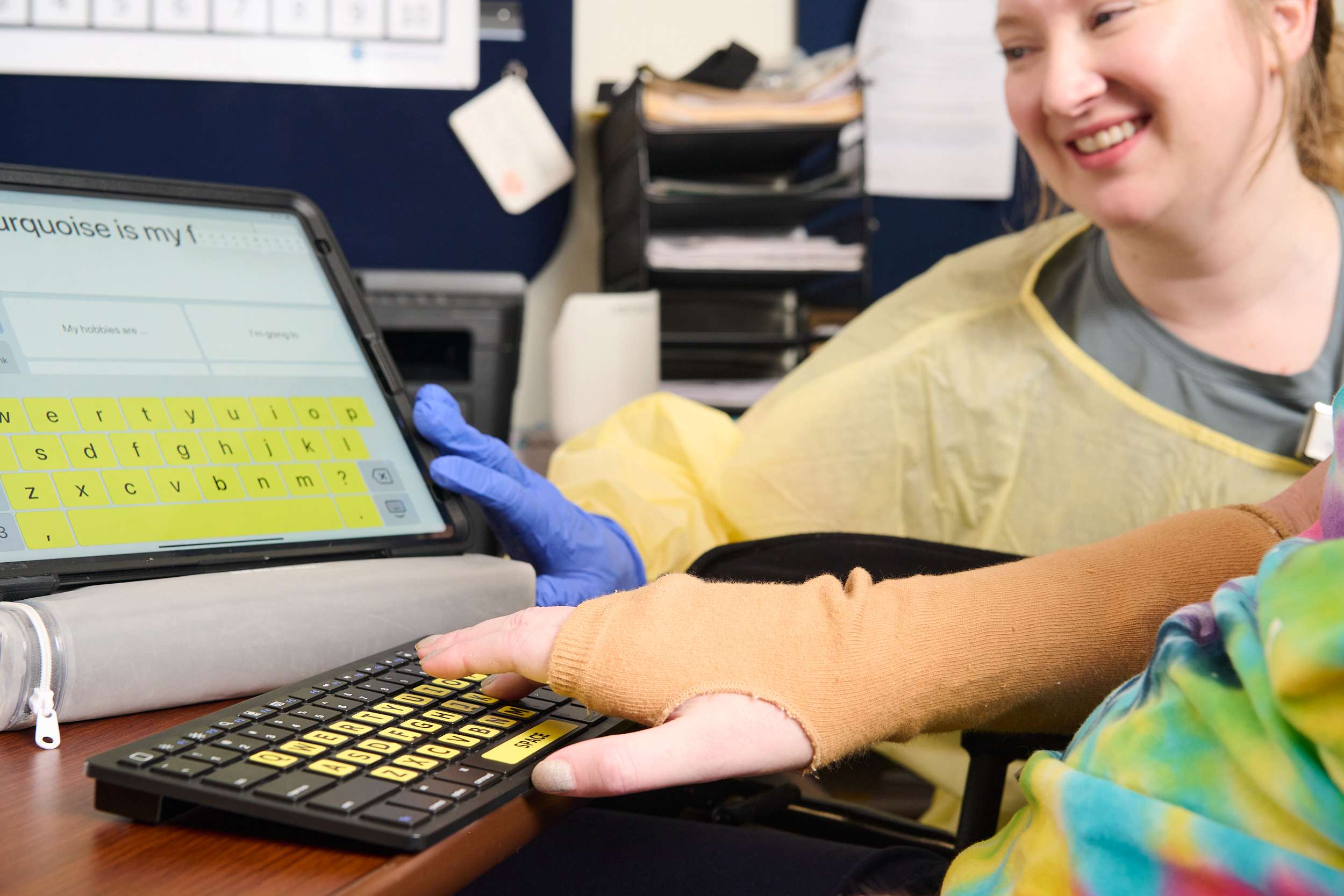 A person in a yellow gown smiles as another person uses a typing device with large keys. A tablet screen displays text, 