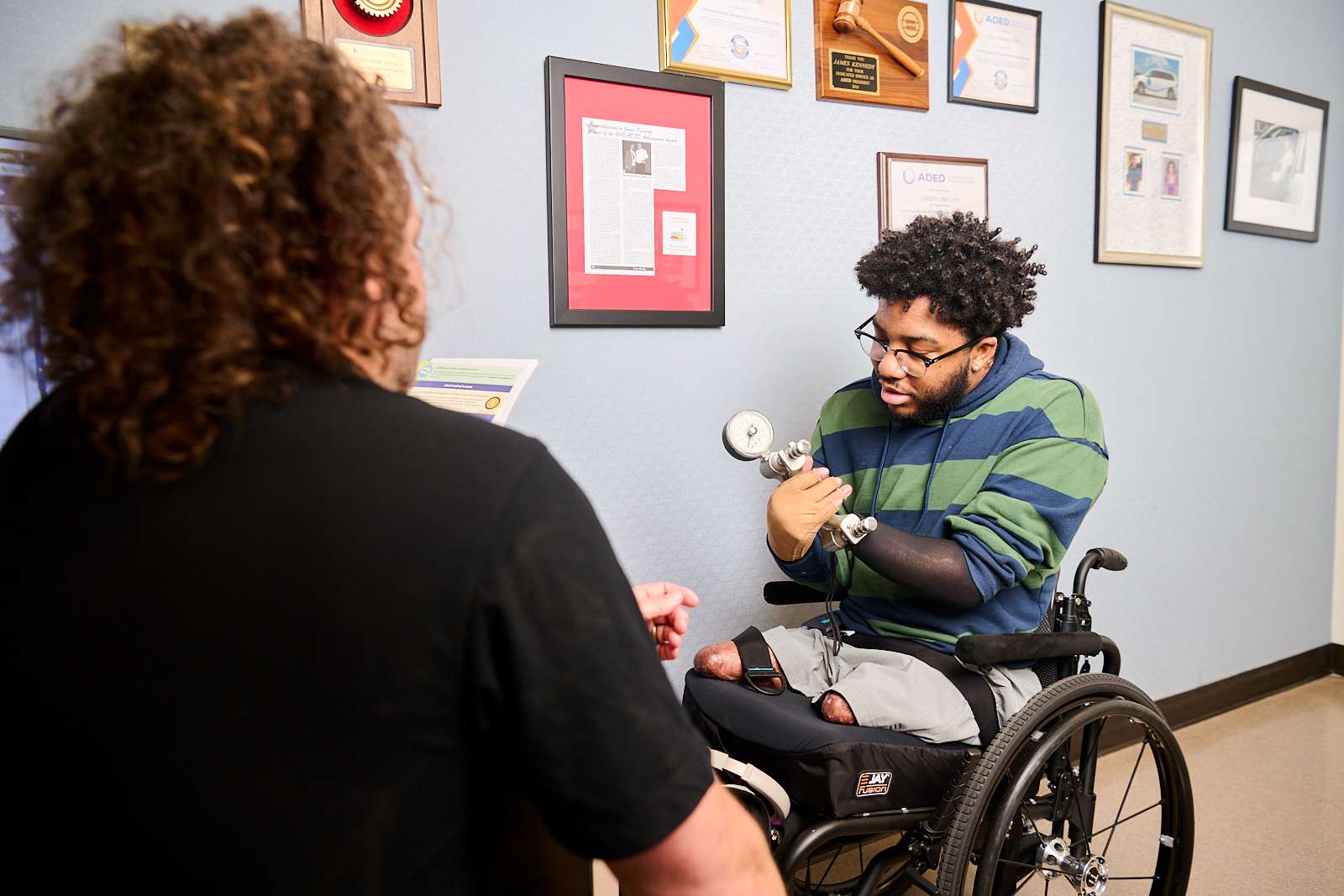 A person using a wheelchair with prosthetic arms and legs is interacting with another person in a room adorned with framed certificates and plaques. They are holding a small device, appearing focused on a task or instruction.