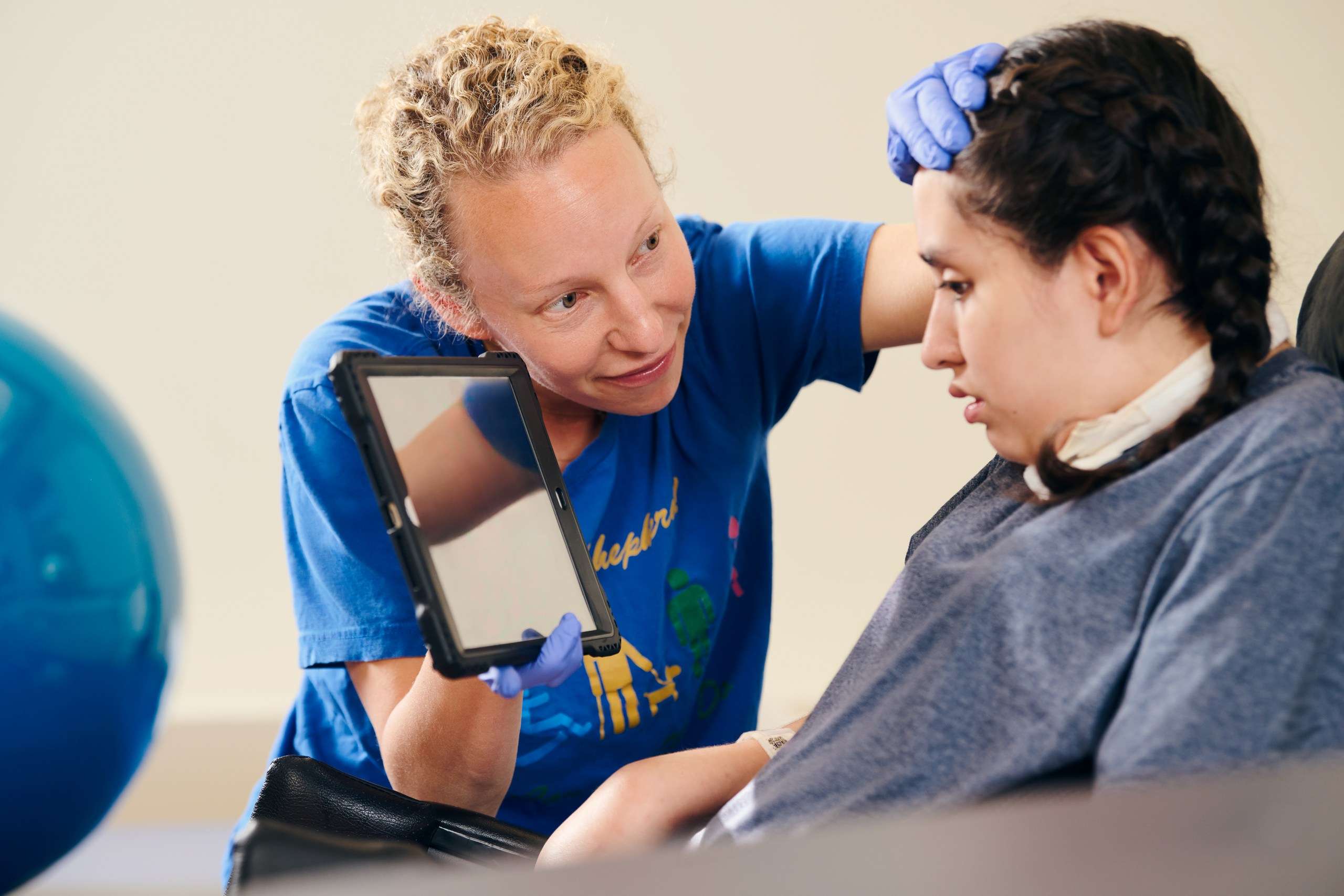 A caregiver with curly hair and wearing gloves is holding a tablet and gently examining a young person’s head. The young individual is seated, wearing a gray shirt and has braided hair. A blue exercise ball is visible in the background.