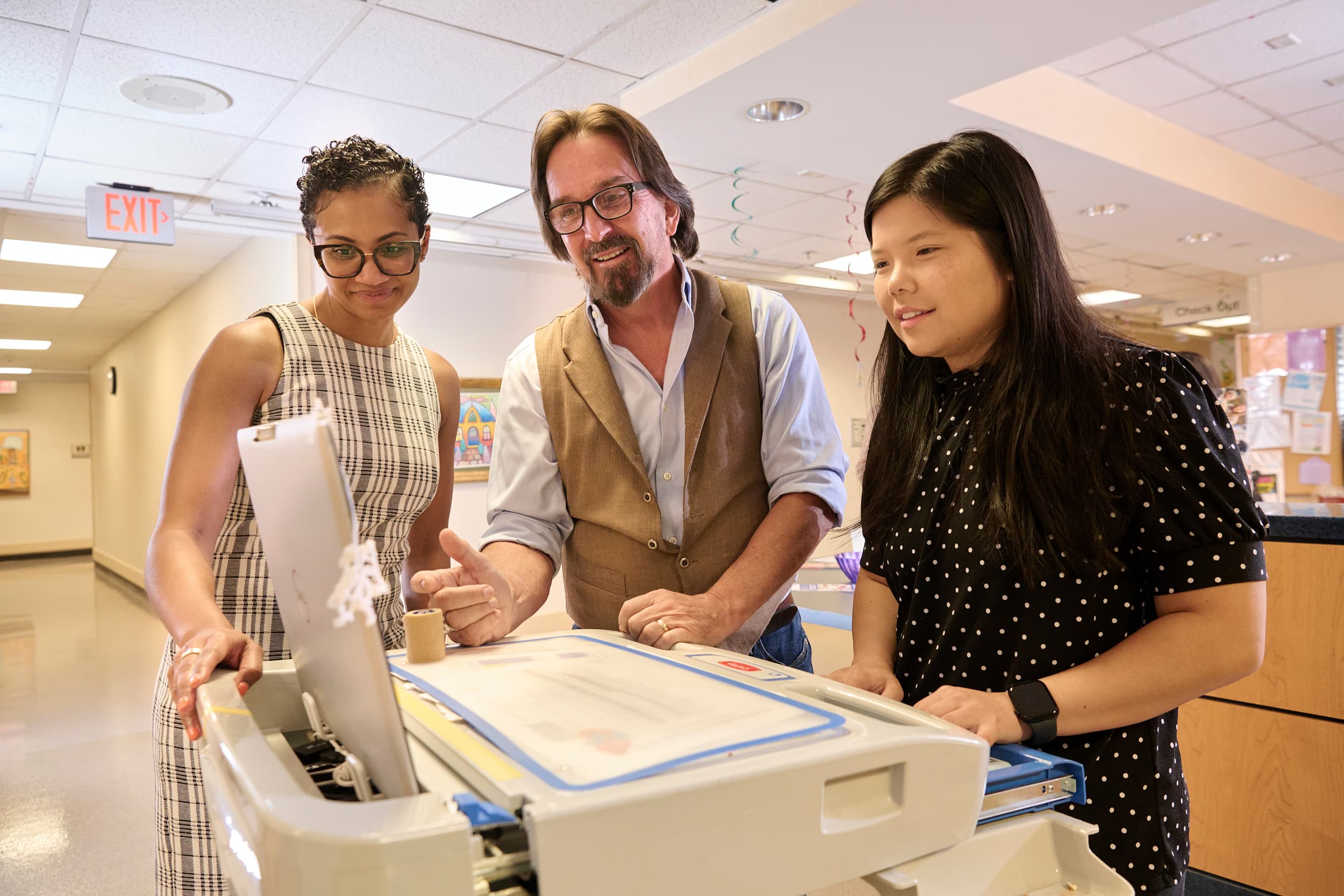Three people stand around a printer in an office setting. One person is pointing at the printer's screen, while the others watch attentively. The office hallway is visible in the background.