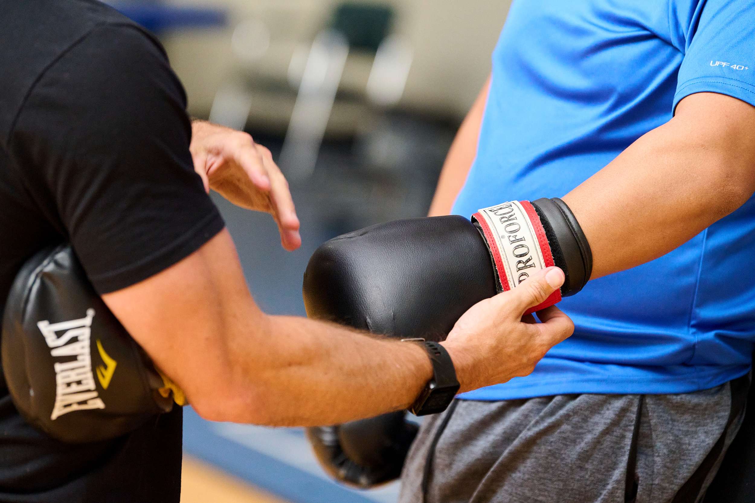 A close-up of a person in a black t-shirt adjusting a black boxing glove on another person's arm. The person wearing the glove is in a blue shirt. The background is slightly blurred, showing a gym setting.