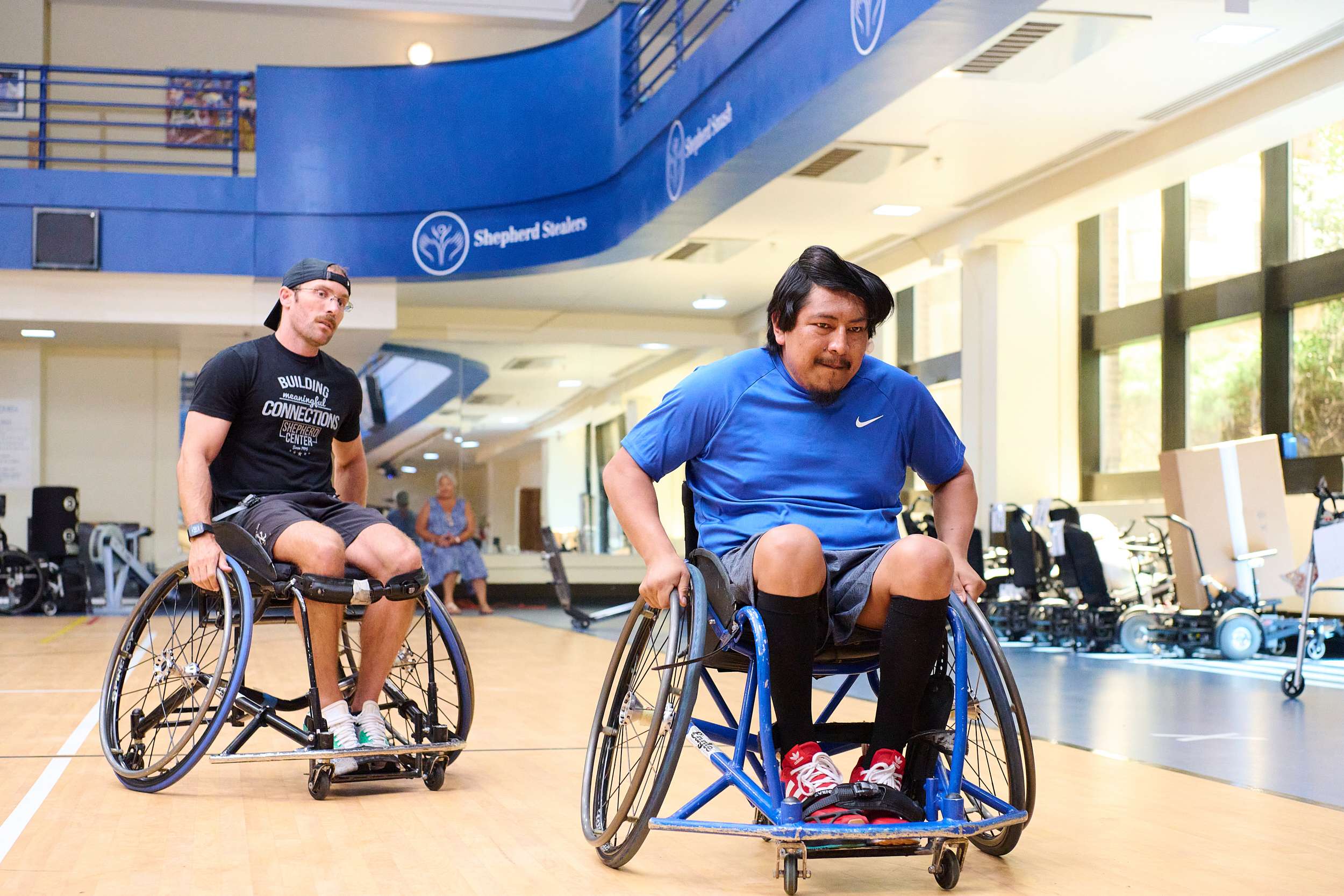 Two athletes in wheelchairs play basketball indoors, embodying comprehensive care for spinal cord injuries. They move quickly on the wooden court, focused under a blue banner displaying the facility's name. Spectators and equipment line the sides, cheering them on.