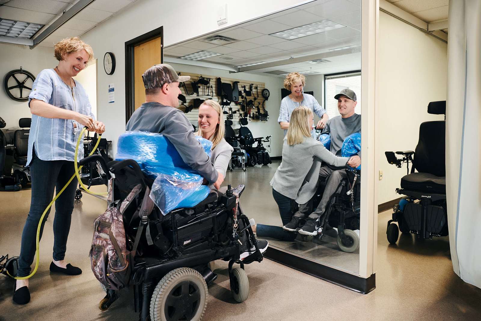 A man in a wheelchair is being fitted with a protective cover by a woman. They are both smiling and looking at a large mirror, reflecting their image. Another person is assisting with a hose. The room contains equipment for mobility assistance.
