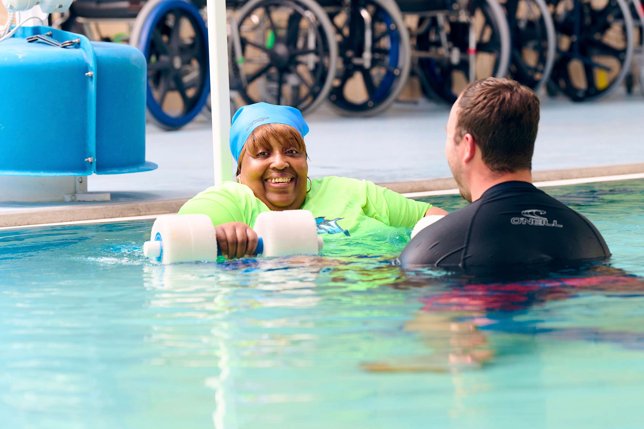 A woman in a blue swim cap and green shirt smiles while holding foam dumbbells in a pool. A man in a black swim shirt stands nearby. Several wheelchairs are visible on the pool deck in the background.