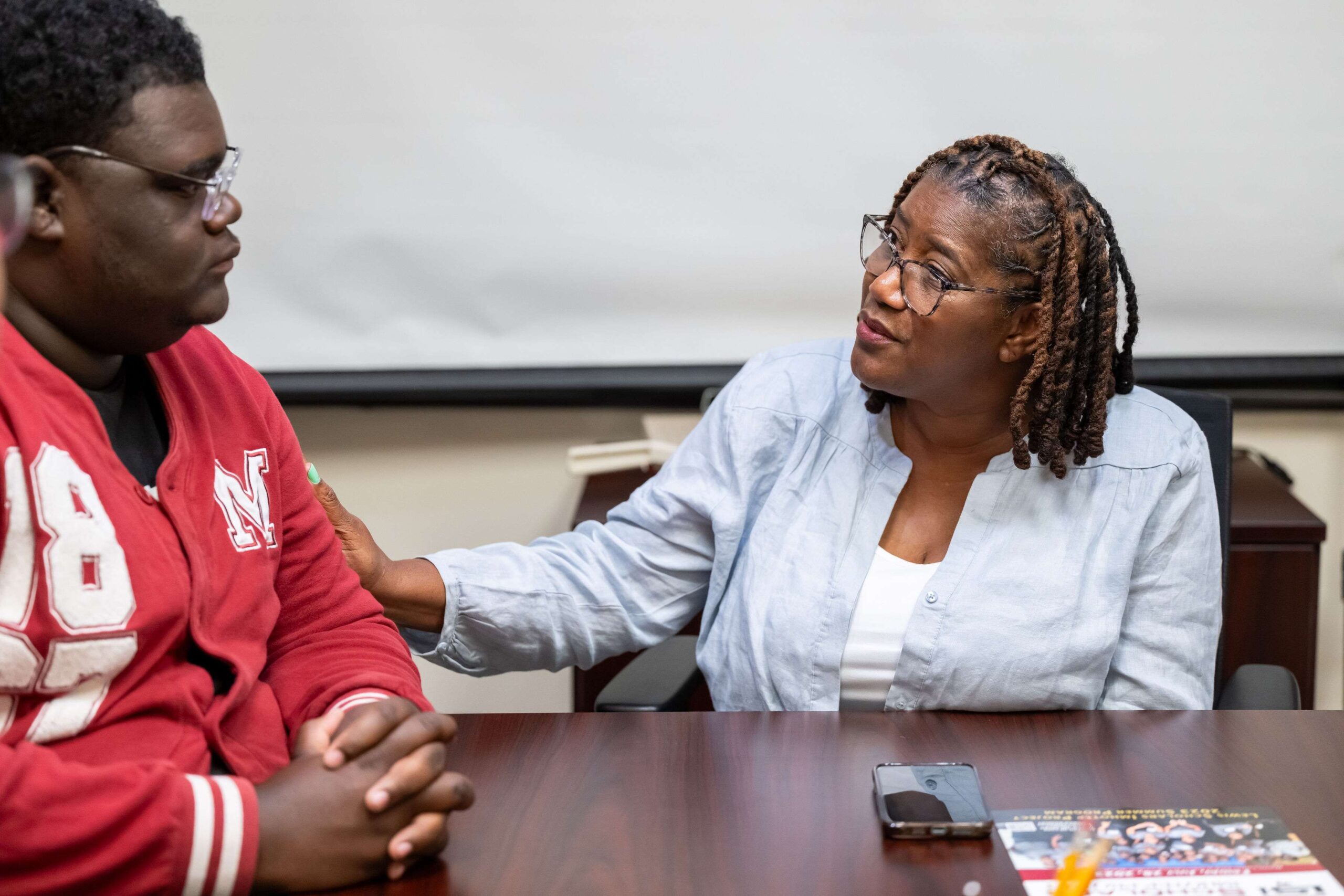 A person wearing a light blue shirt sits at a table, speaking to another person in a red jacket, offering a comforting gesture. A smartphone and magazine are on the table.