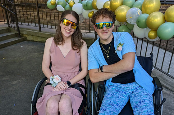 Two people wearing reflective sunglasses smile while sitting in wheelchairs. The person on the left wears a pink dress, and the one on the right is in a blue shirt and patterned pants. They are in front of a balloon display with gold, green, and white balloons.