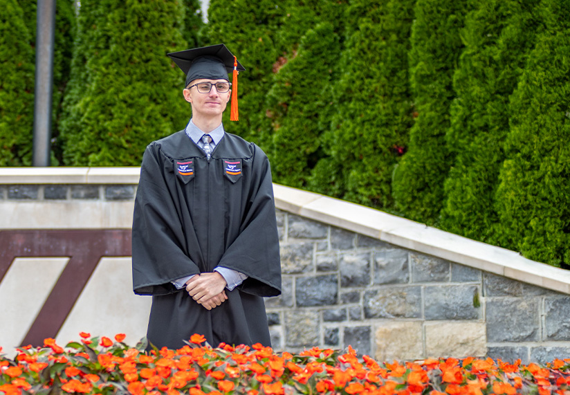 A person in a graduation gown and cap stands in front of a stone sign with 