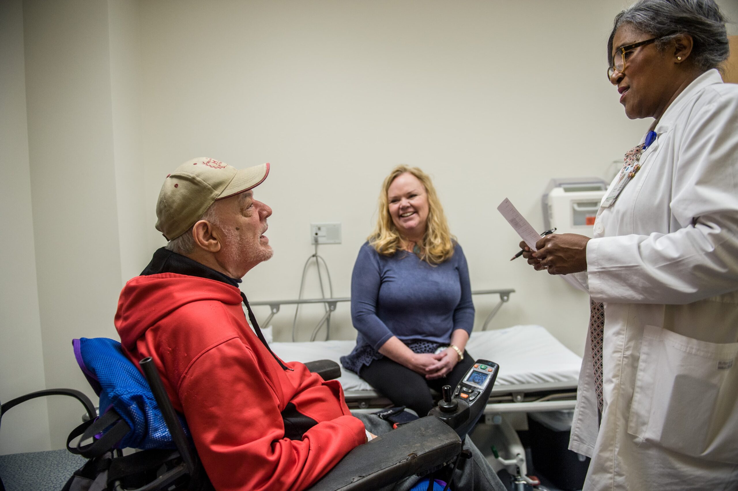 A man in a wheelchair wearing a red jacket discusses neurourology with a healthcare professional in a white coat holding a paper. Meanwhile, a woman smiles from the examination table, adding warmth to the bustling medical office.