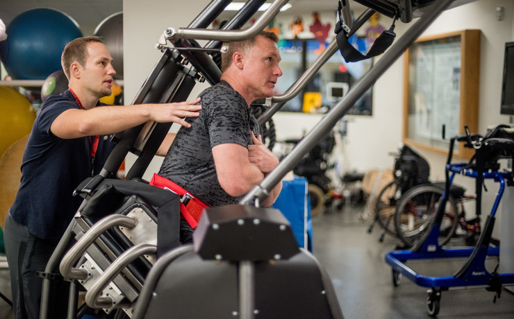 A physical therapist assists a man using a specialized walking machine in a rehabilitation center. The room contains exercise equipment and therapy tools, with exercise balls and wheelchairs visible in the background.