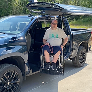 A man in a wheelchair is being lifted out of a black Chevrolet Silverado with a side-entry ramp. He is wearing a light-colored T-shirt, shorts, and glasses. The truck is parked on a sunlit driveway with green foliage in the background.