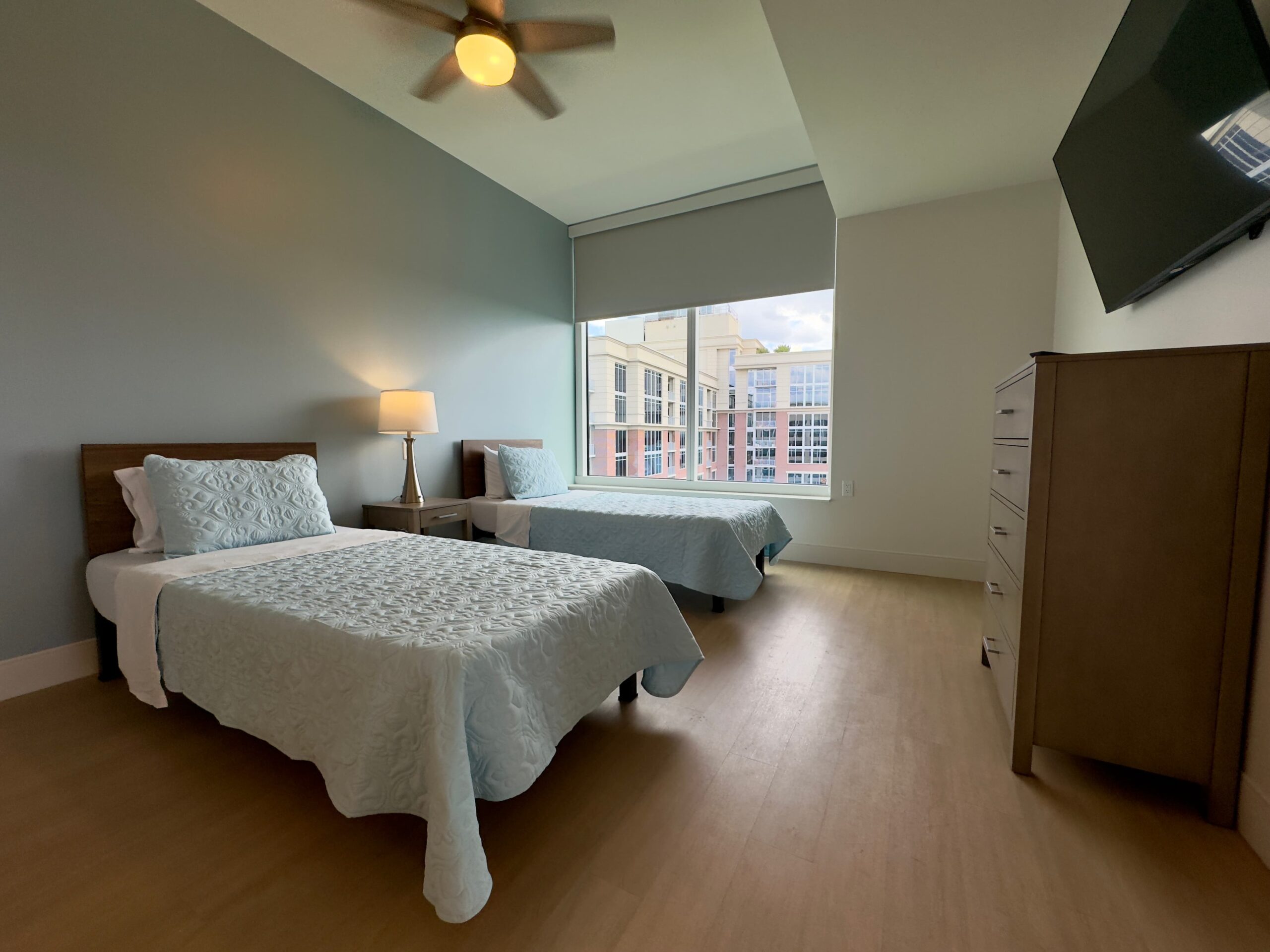 Bedroom with two single beds, teal bedding, and pillows. A wooden nightstand with a lamp is between the beds. Large window shows city buildings. Ceiling fan above and a flat-screen TV on the wall. Light wood floors complete the space.