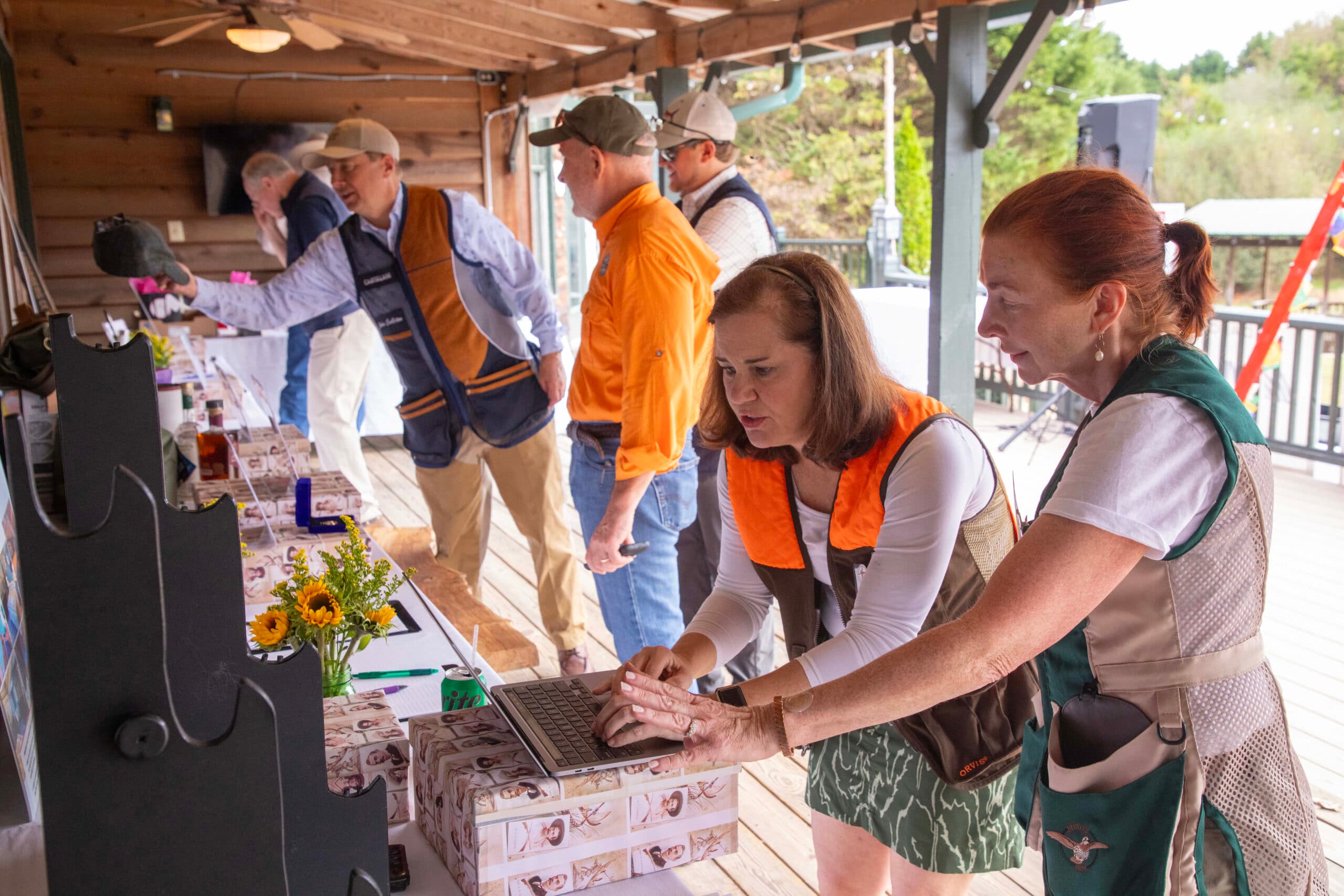 People in shooting vests participate in an auction on a porch. Two women focus on a laptop surrounded by flower arrangements and shelves of items. Others observe or write notes. The atmosphere is casual and involved.