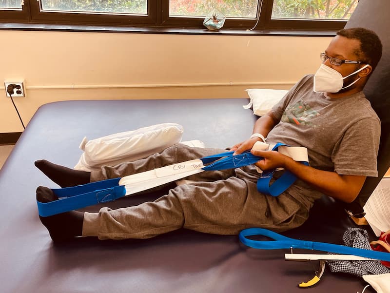 A person wearing a mask and glasses is seated on a therapy bed, using a blue strap to exercise their legs. They are dressed in casual clothing, and there is a pillow next to them. Large windows are visible in the background.