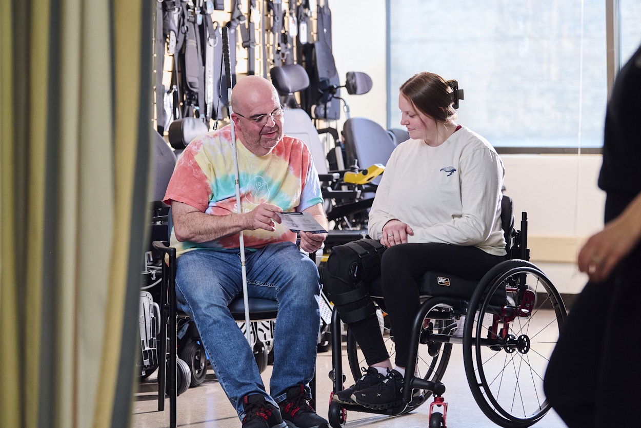 A man in a tie-dye shirt and a woman in a wheelchair share a light moment in a bright room. The man is holding a piece of paper. There's exercise equipment in the background.