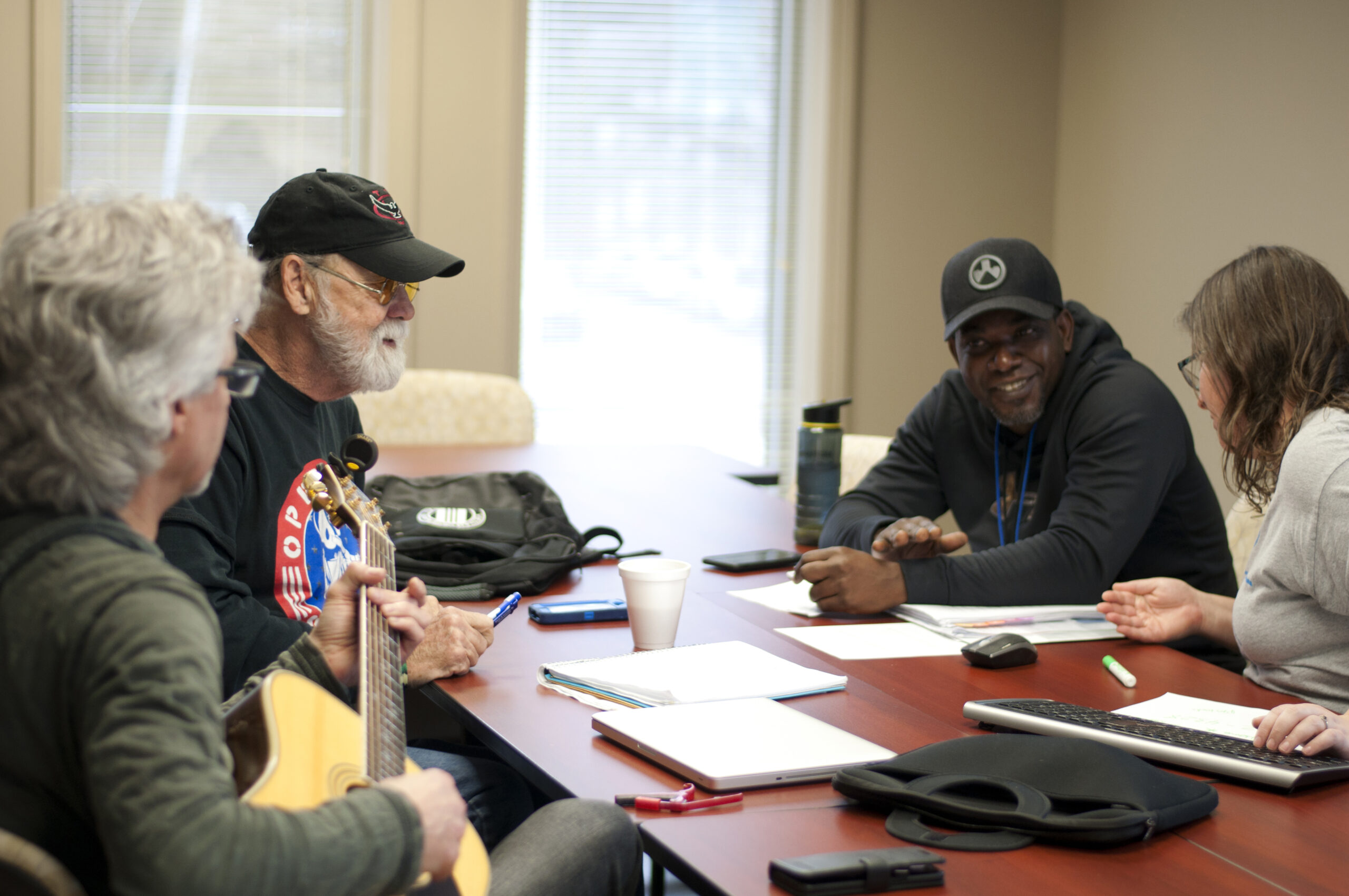 Four people sit around a conference table engaged in conversation. One person holds a guitar. Papers, a cup, and a keyboard are on the table. They are inside a room with large windows in the background.