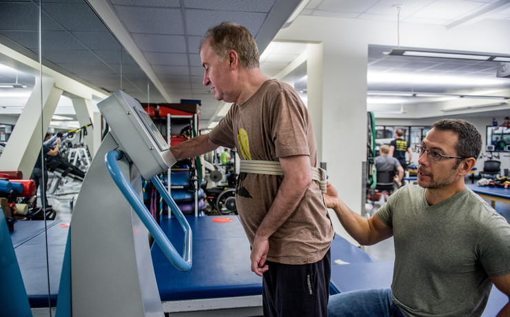 A man is using a treadmill in a rehabilitation center with the support of a therapist who is kneeling beside him. The man is wearing a harness, and there are mirrors and exercise equipment in the background.