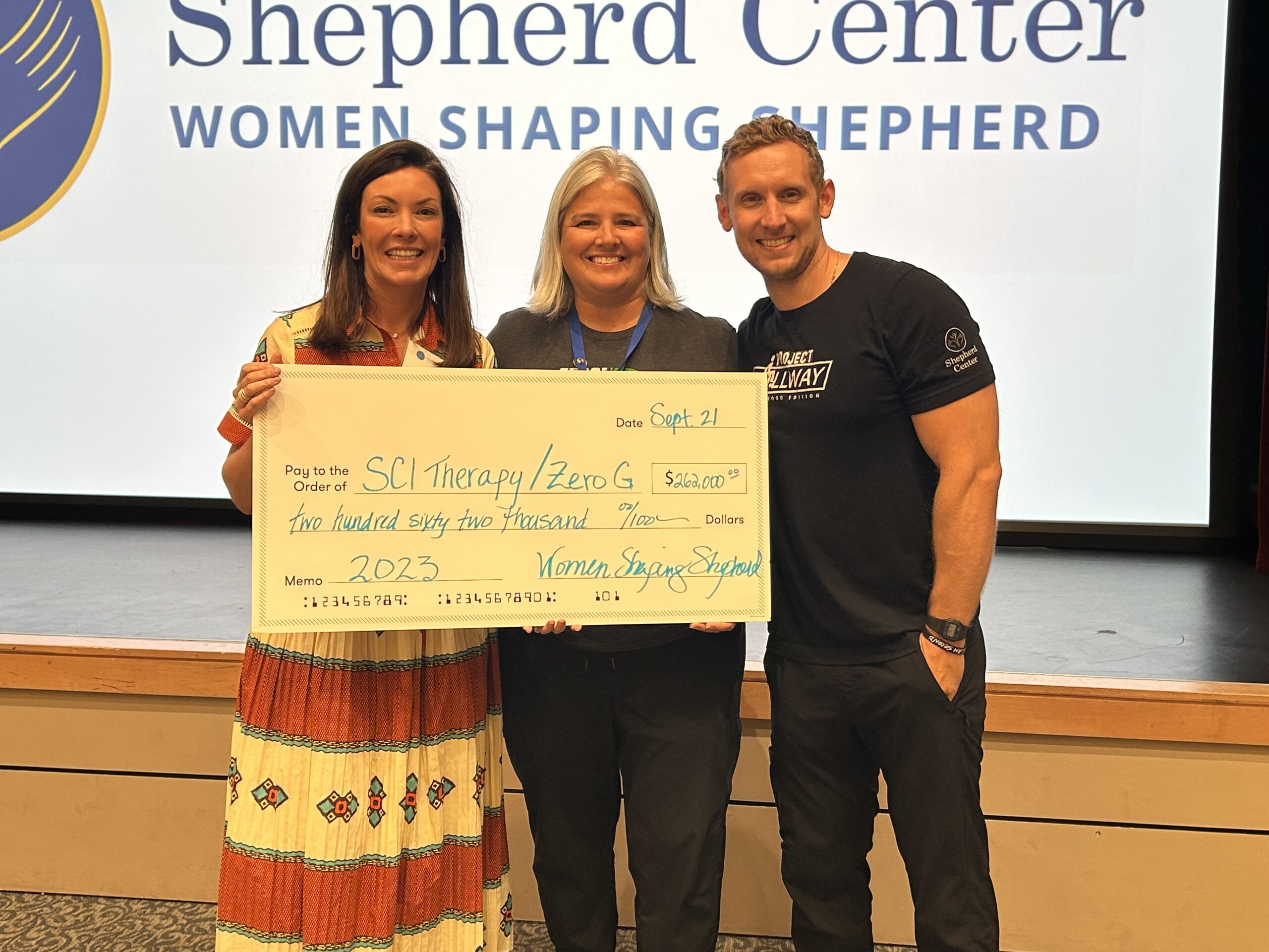 Three people are standing on a stage holding a large ceremonial check. They are smiling, and the background shows a screen with the text 