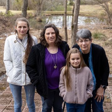 Four people stand outdoors on a stone path. Two adults and a young girl are in front, with a young woman standing slightly behind. They're dressed in jackets and sweaters, and trees with a pond in the background create a natural setting.