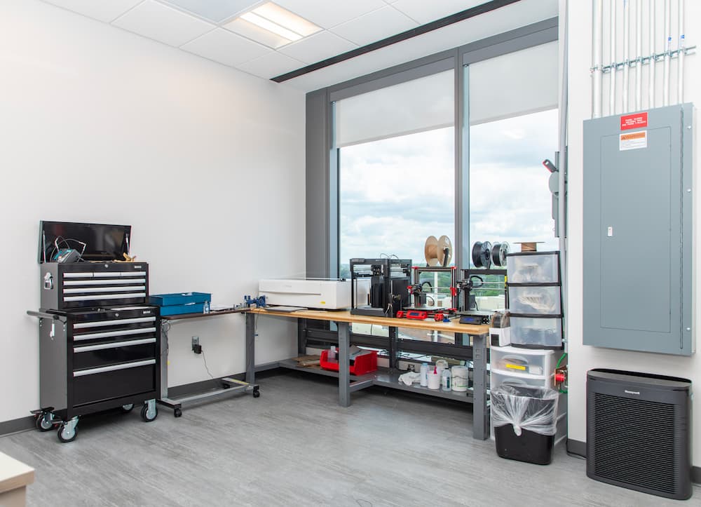 A modern workshop with a tool chest, workbench, storage bins, computer monitor, spools of filament, and large windows letting in natural light. Electrical panels and air purifier are also visible.