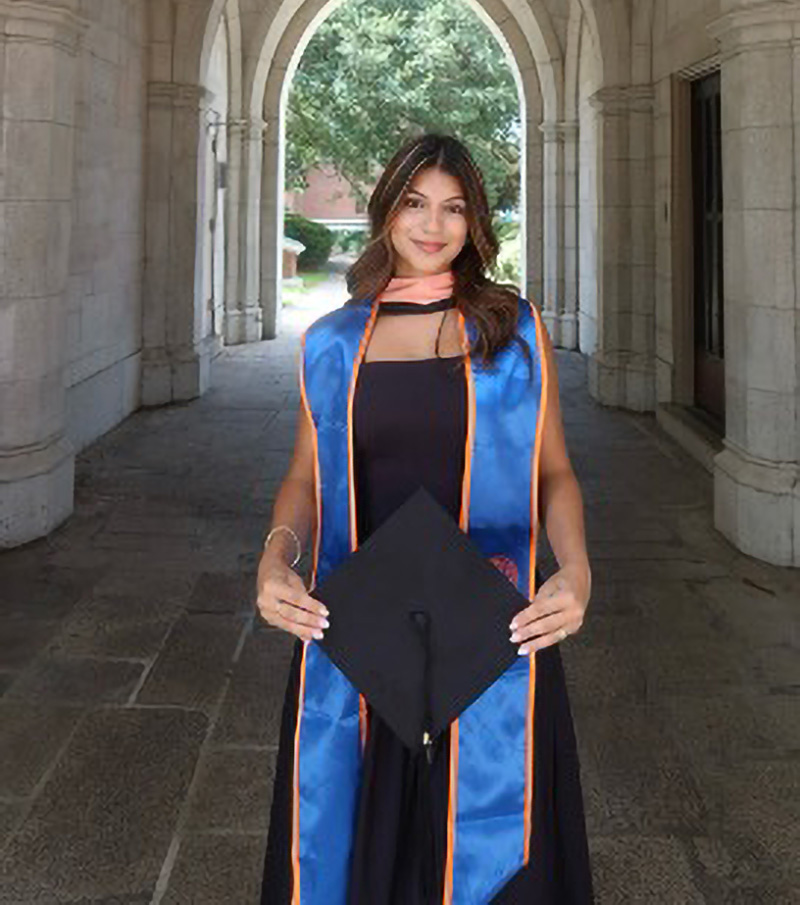 Andrea Ferreira, wearing a graduation stole and holding a cap, stands in an arched stone hallway, smiling at the camera. Sunlight and greenery are visible at the end of the hallway.