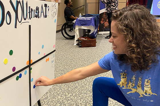A woman in a blue shirt places a blue sticker on a board with colored dots in a grid. Other people and a wheelchair are visible in the background at what appears to be an indoor event or meeting.