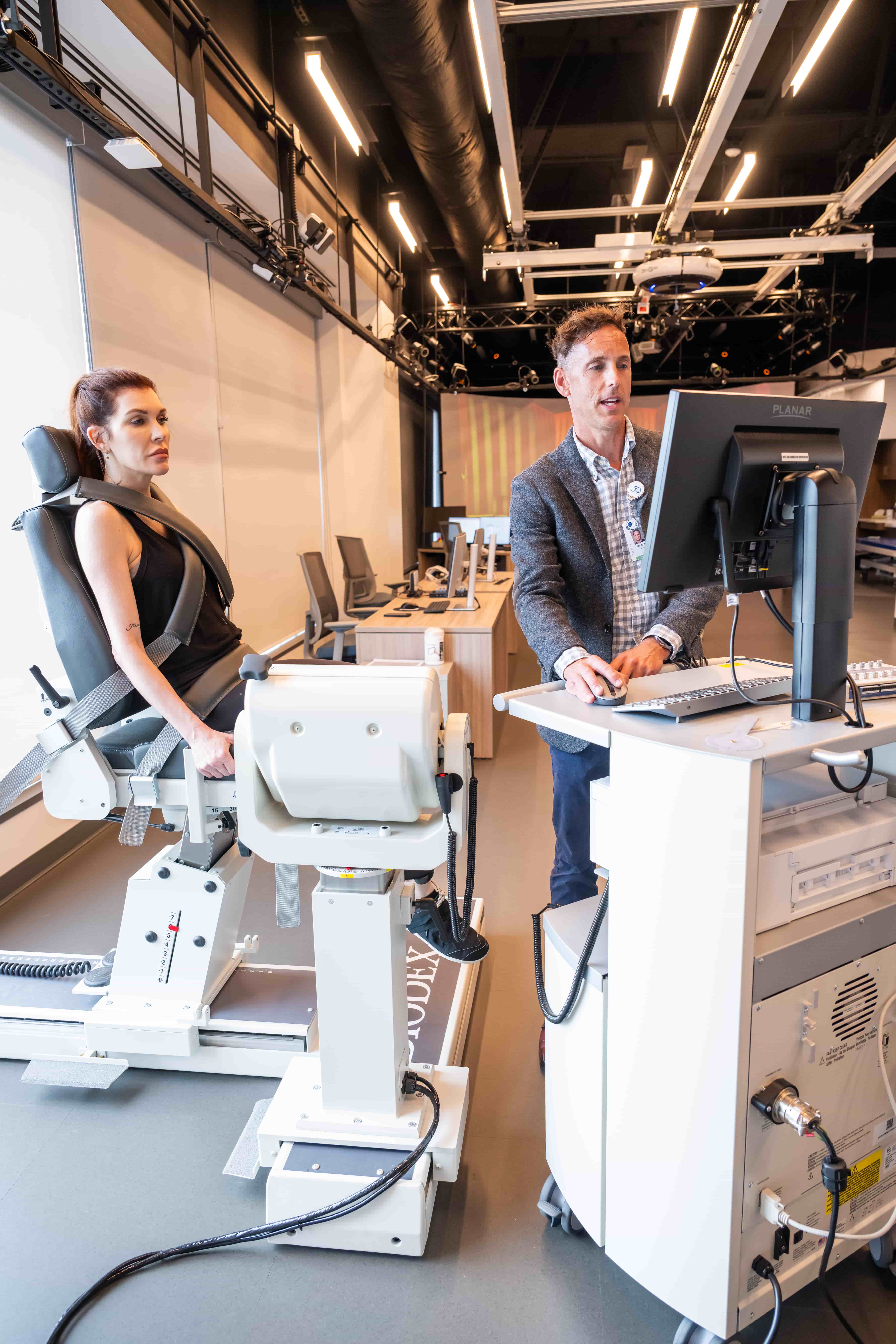 A woman sits on a medical testing machine while a man in business attire operates a computer nearby in a modern, well-lit lab or clinic setting.
