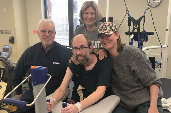 Four people are gathered in a rehabilitation setting. A man with glasses and a woman stand behind a seated man who is using medical equipment. Another woman in a cap and sweater is crouched beside him, smiling. Medical equipment is visible in the background.