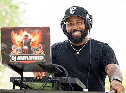 A smiling man wearing headphones and a black cap stands behind DJ equipment outdoors. His laptop displays a colorful graphic with the text 