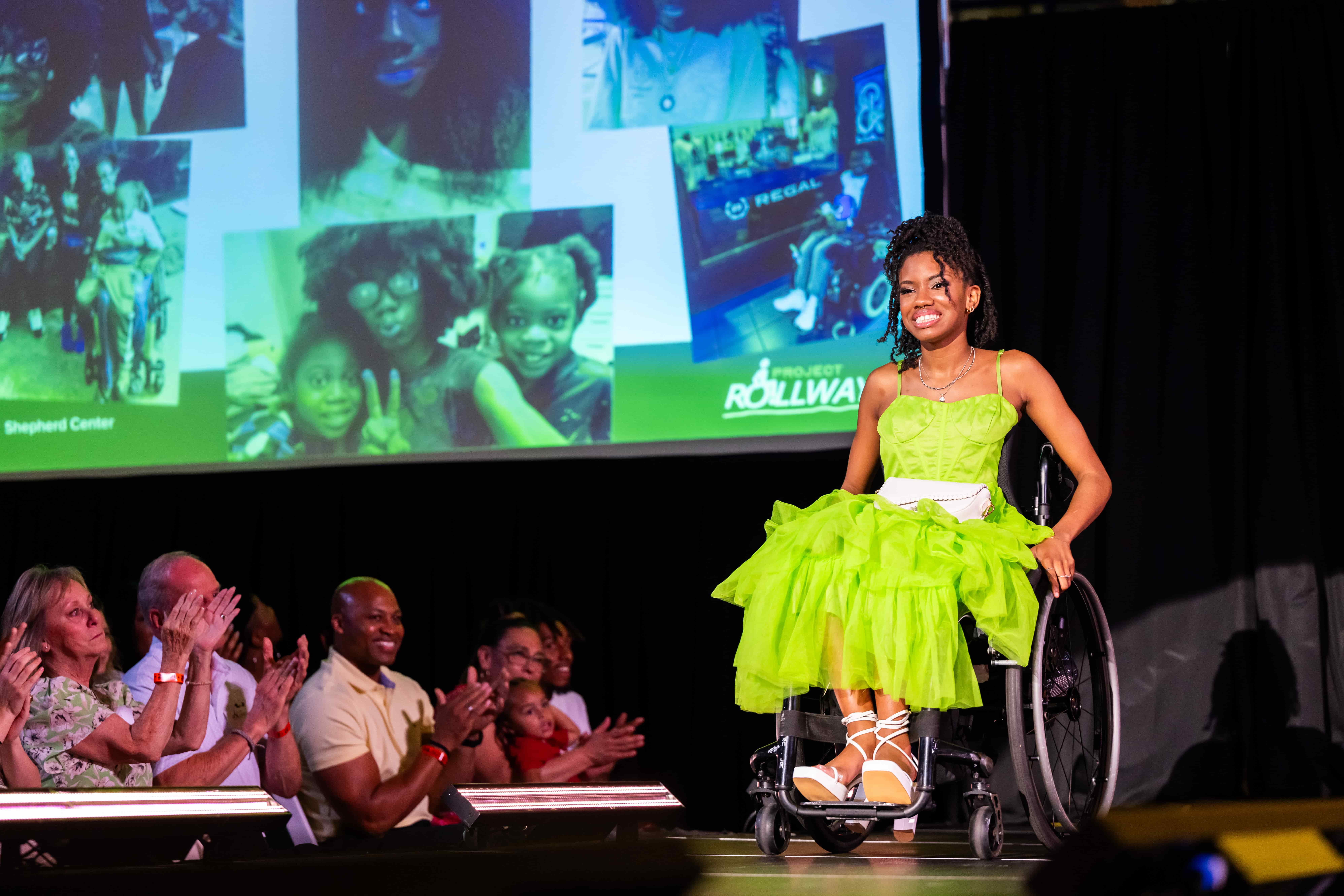 A young woman in a bright green dress smiles while using a wheelchair on stage. The audience applauds, and a projection of photos is visible behind her.