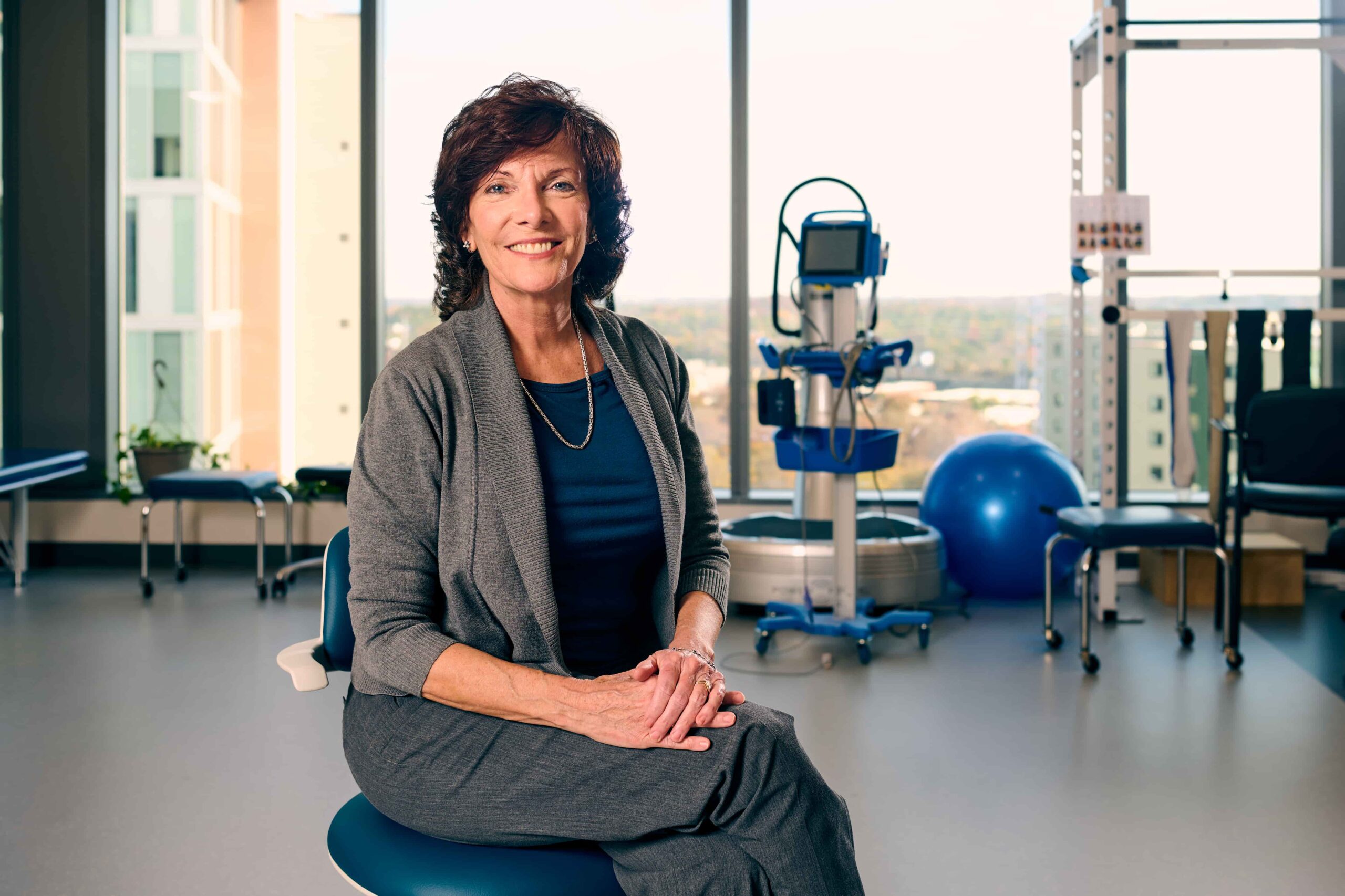 A woman with short brown hair, wearing a gray suit and navy top, sits smiling on a stool in a bright, modern physical therapy or medical office with exercise equipment in the background.