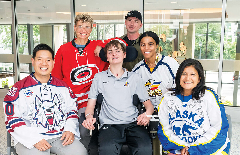 Six smiling people pose indoors, including a young man in a wheelchair at the center. The group wears various hockey jerseys and casual clothes, and they appear happy and supportive in a bright, modern setting.