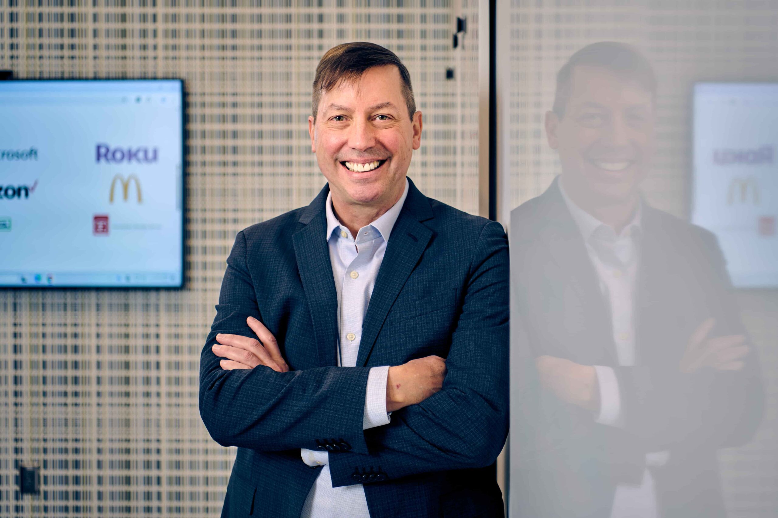 A man in a dark suit stands with arms crossed and smiles at the camera, with his reflection visible on a glass wall beside him and a monitor displaying various company logos in the background.