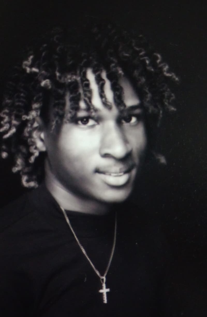 Kaedan Everhart wears a dark shirt and a necklace with a cross pendant, looks slightly past the camera, and smiles softly.