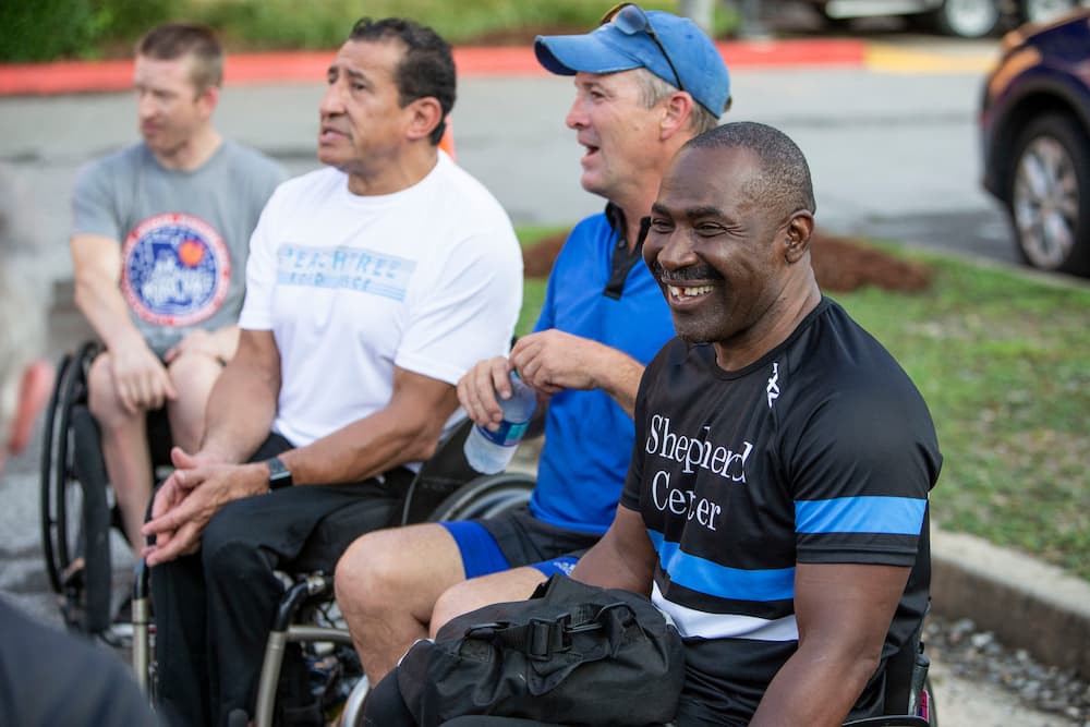 Four men in wheelchairs are outdoors, smiling, and engaged in conversation.