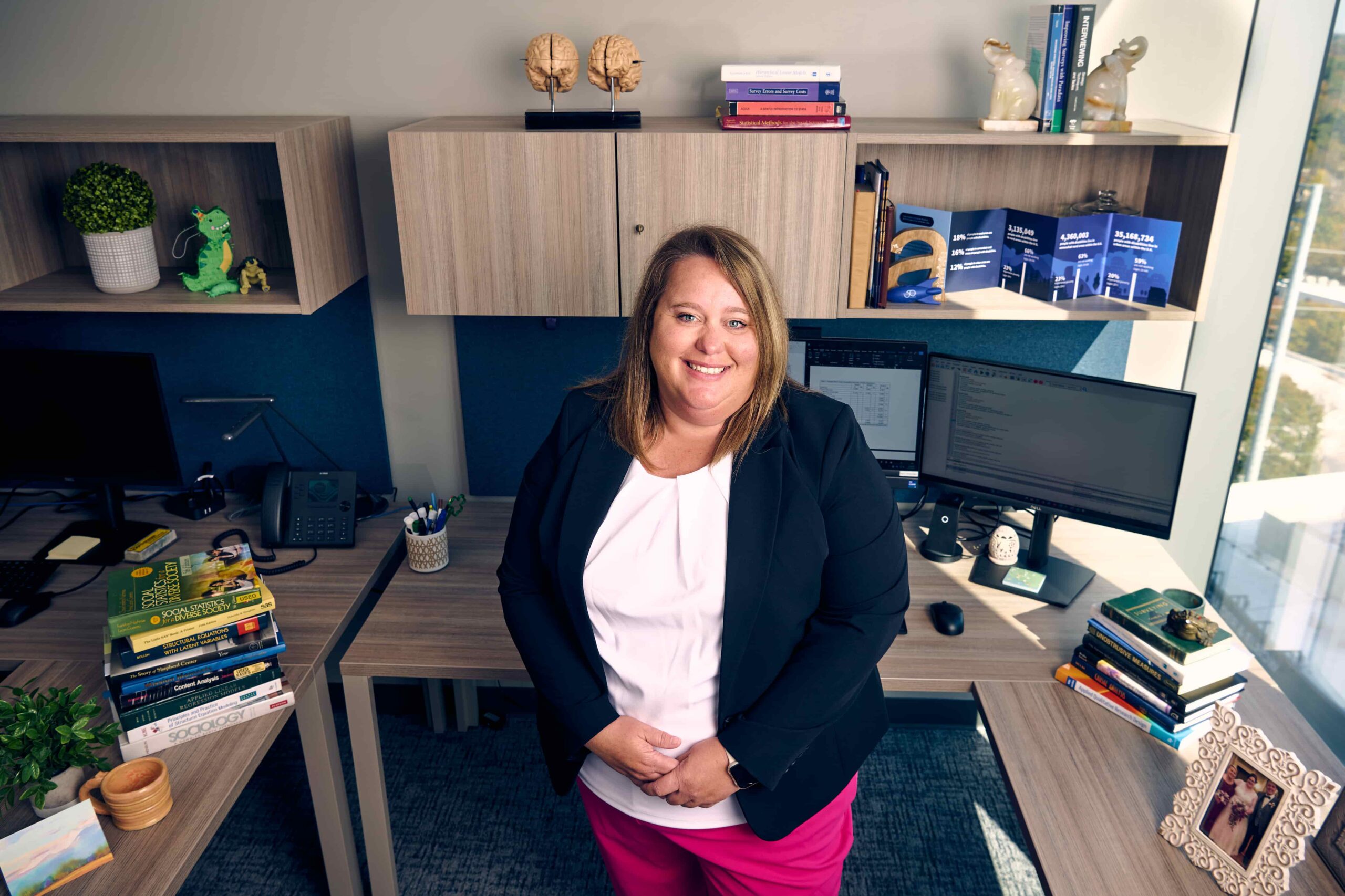 A woman in a black blazer and pink pants stands smiling in a modern office with desks, computer monitors, books, plants, and decorative items, including brain models and awards, visible around her.