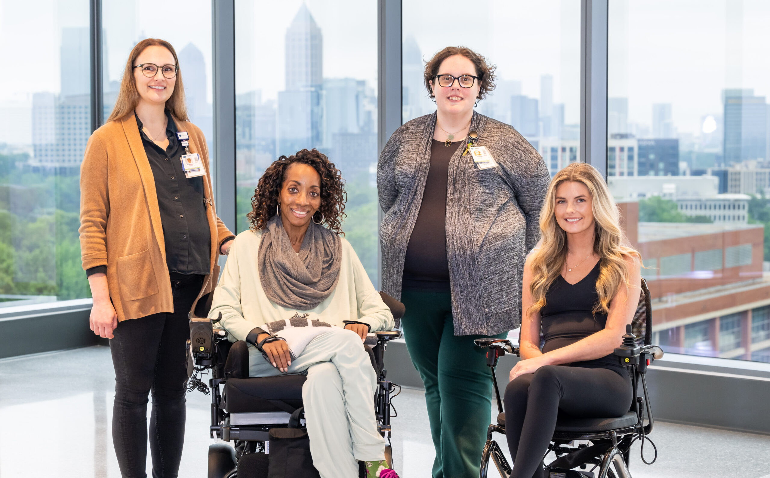 Four women pose together indoors by large windows; two are standing and two are seated in wheelchairs. A city skyline is visible in the background.
