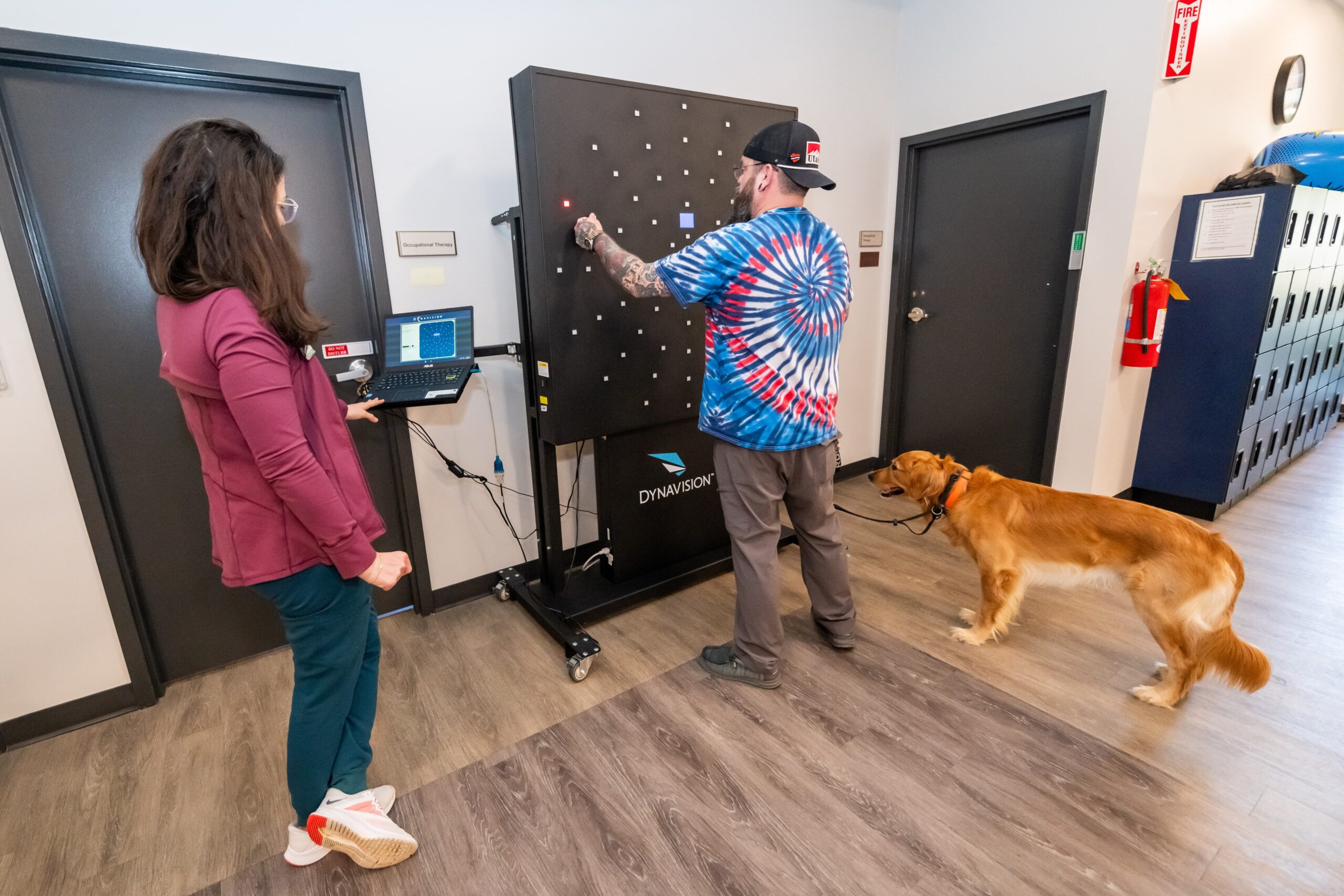 A man uses a light board for a test while a woman observes with a laptop. A golden retriever wearing a harness stands beside the man in a hallway with lockers and doors.