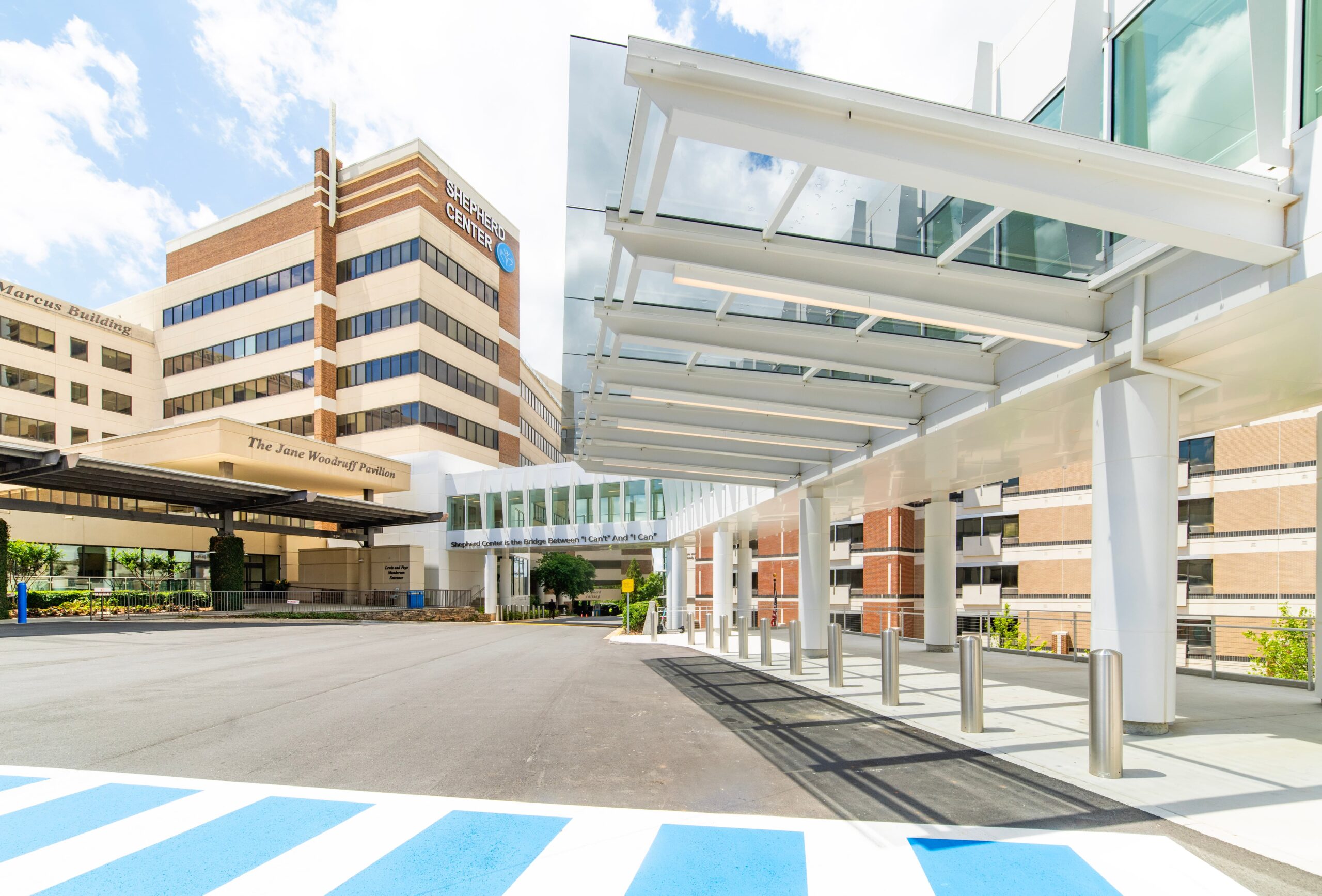Exterior view of a modern hospital complex with multiple brick and glass buildings, a covered entrance, pedestrian bridge, and blue sky with scattered clouds. The foreground shows a driveway and striped crosswalk.
