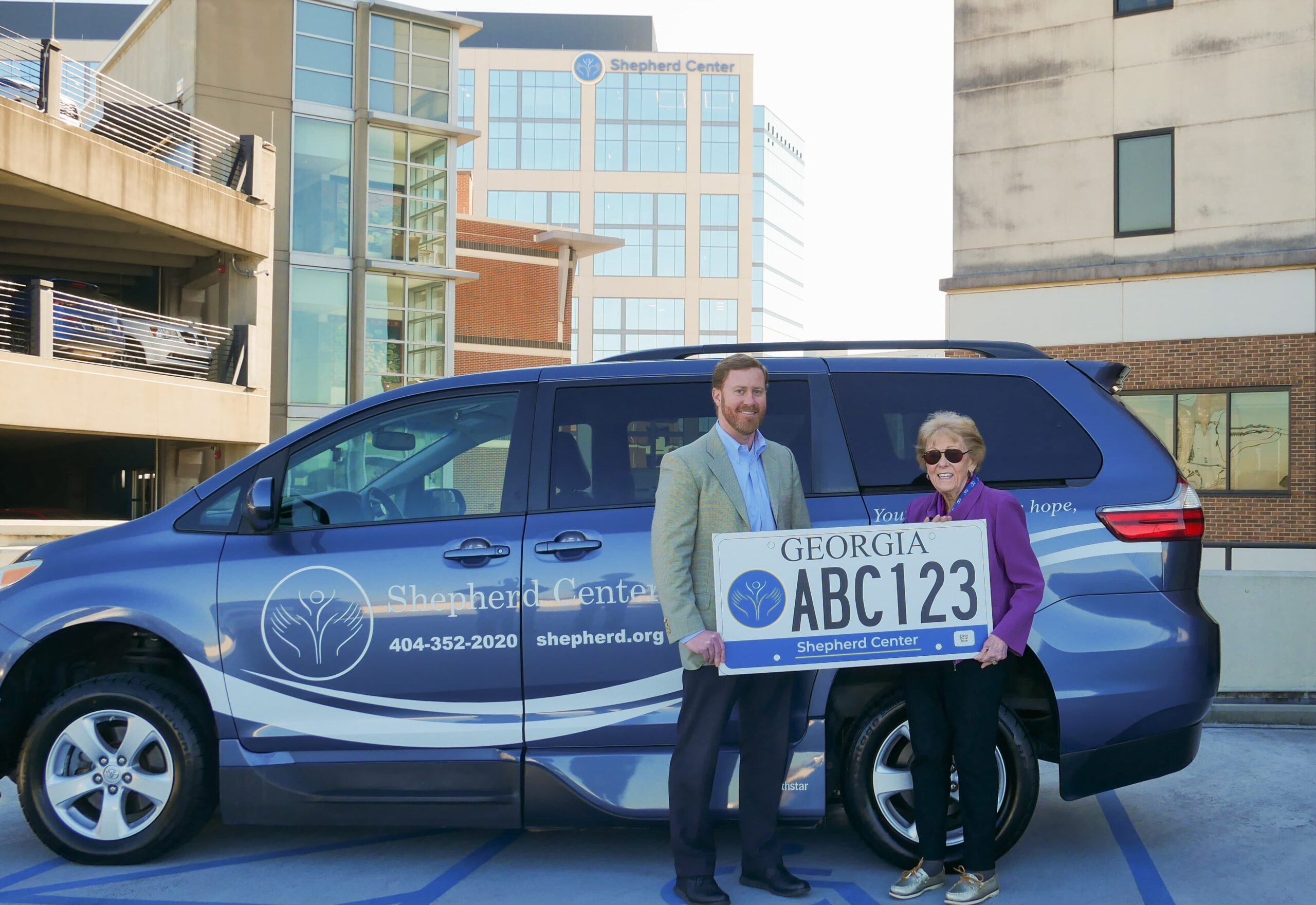Jamie Shepherd and Alana Shepherd stand in front of a Shepherd Center van holding a large mock Georgia license plate. The Shepherd Center building is visible in the background on a sunny day.