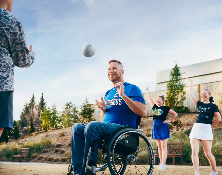A man in a wheelchair, wearing a blue sports jersey, smiles while tossing a ball with a child. Two cheerleaders stand behind them, raising their arms, outdoors on a sunny day.